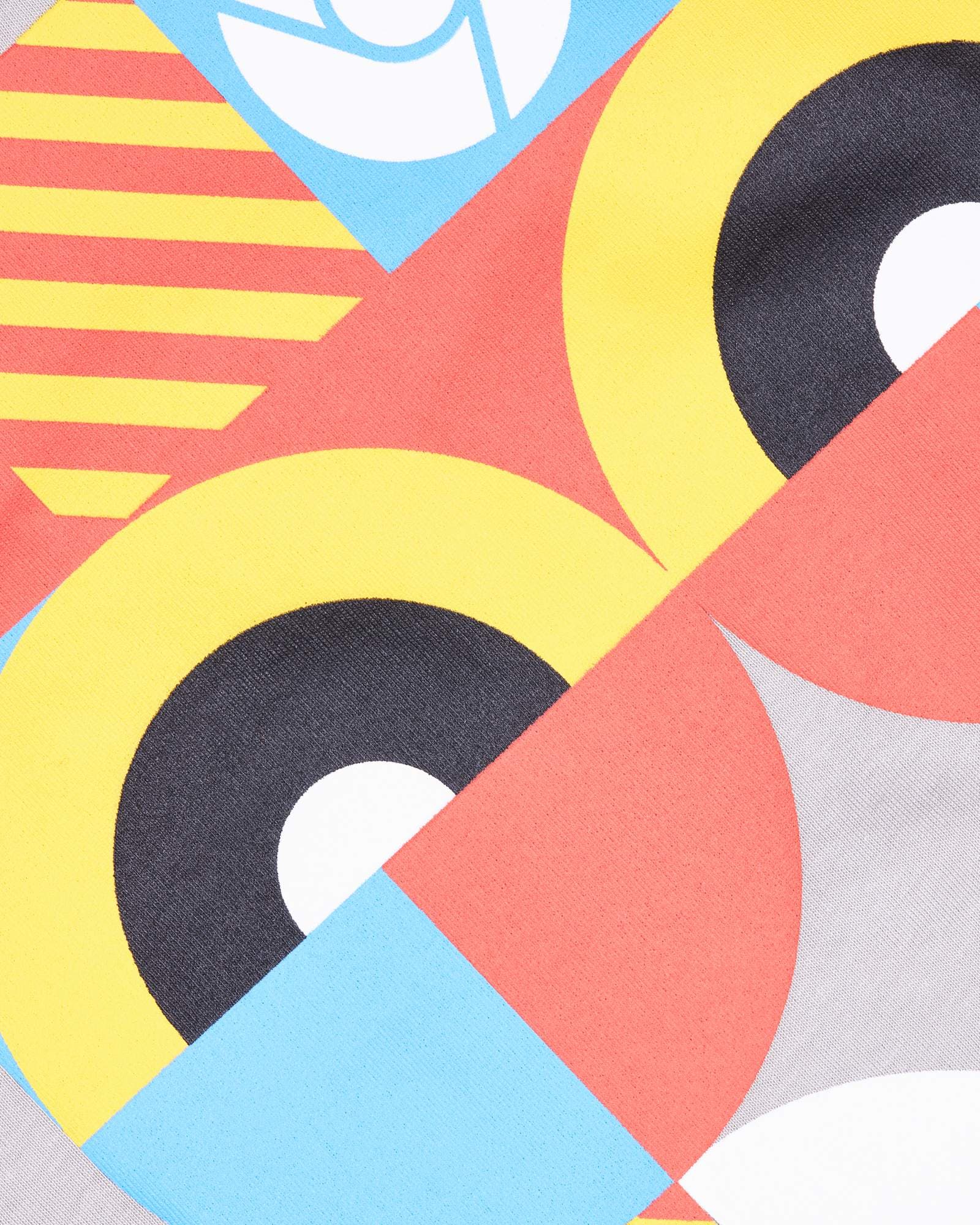 Close-up of a print on light grey fabric: yellow and black rings with white centres, red and yellow stripes, and red and light blue triangles, with part of a round white logo in a light blue diamond at the top edge.