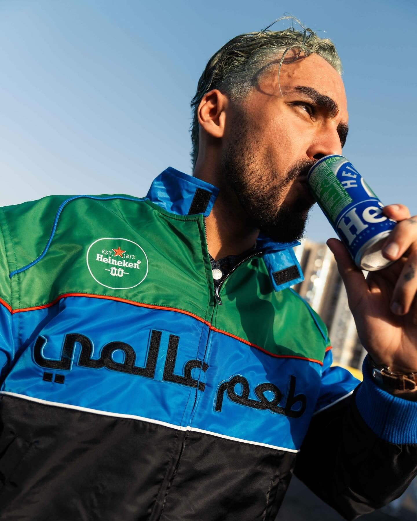 A person drinking from a blue Heineken can, in a green, blue and black Heineken 0.0 racing jacket with black Arabic lettering and a round Heineken 0.0 badge, against a blue sky.