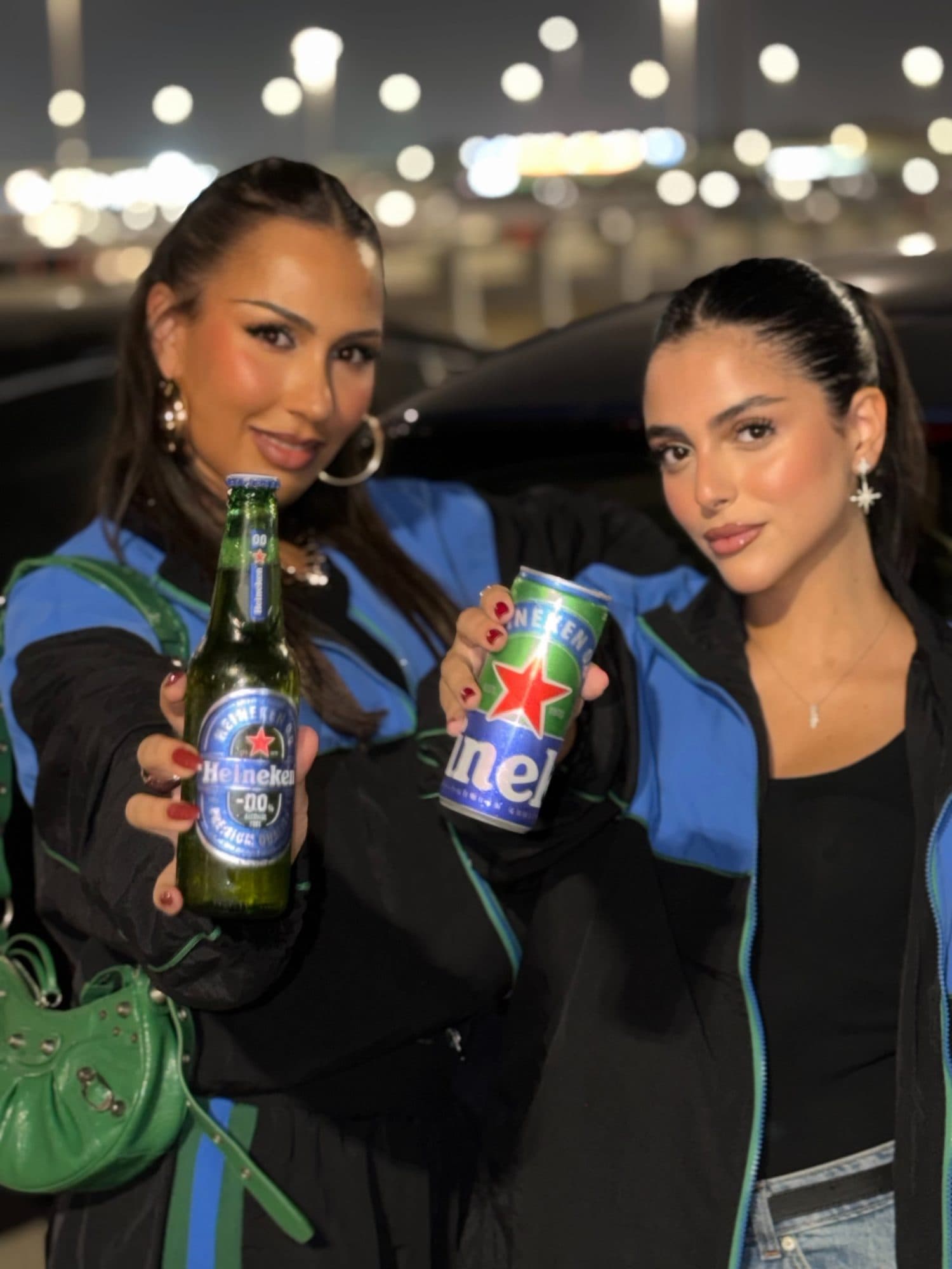 Two people at night holding out a bottle of Heineken 0.0 and a Heineken can, in black track jackets with blue shoulders and green piping.