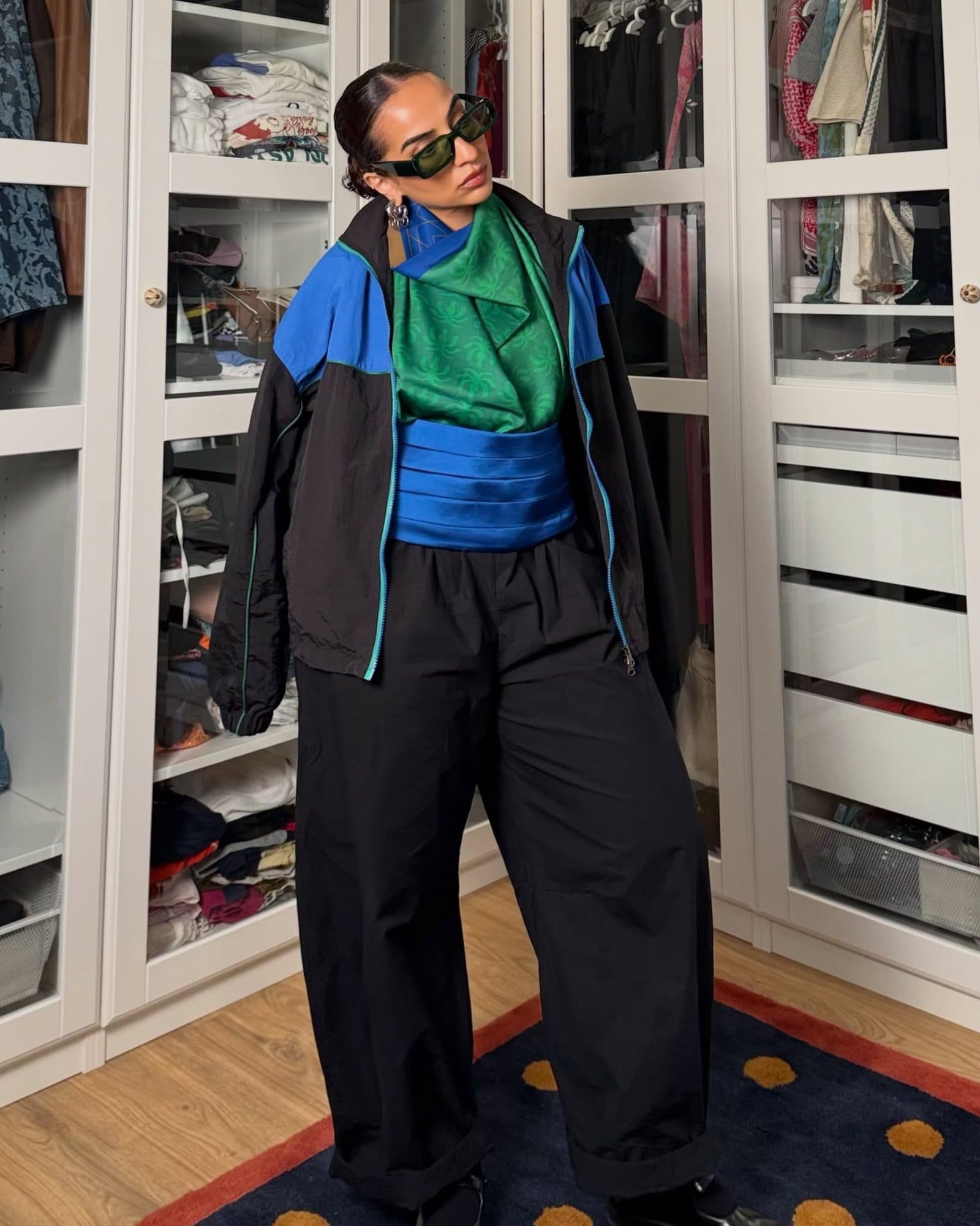A person in sunglasses standing in front of glass wardrobes, in a black track jacket with blue shoulders and teal zips over a green patterned scarf, a blue sash wrapped around the waist and wide black track pants.