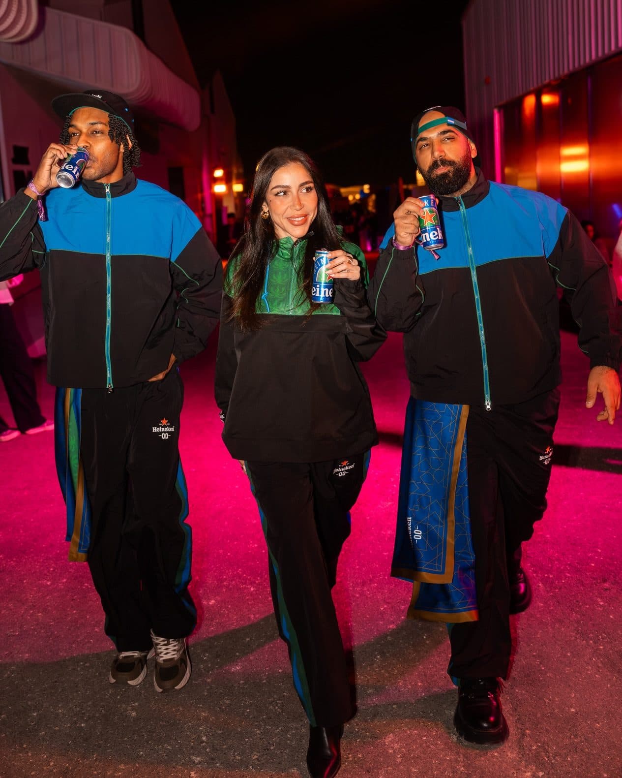 Three people with Heineken cans walking down a pink-lit street at night, two in black and blue track jackets and one in a green and black top, all in black track pants with the Heineken 0.0 logo and blue and green side stripes; one wears a blue scarf with a copper geometric pattern at the hip.