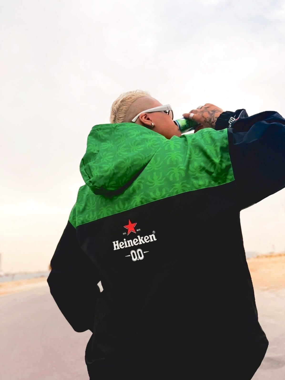 A person seen from behind drinking from a can, in a windbreaker with a green patterned hood and shoulders and a black body with the white Heineken 0.0 logo and red star on the back.