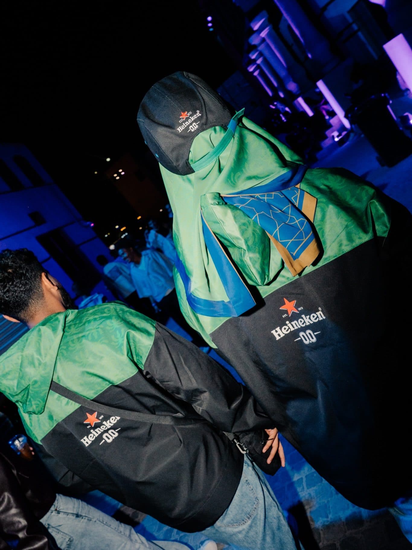 Two people seen from behind at night in green and black windbreakers with the Heineken 0.0 logo on the back, one in a black Heineken 0.0 cap with a blue patterned scarf over the shoulders.