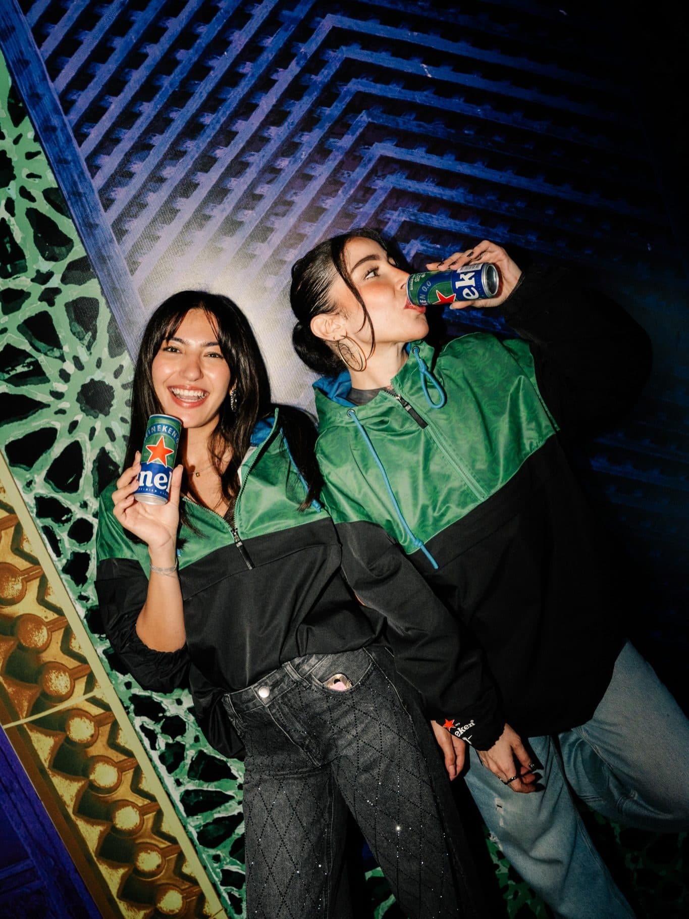 Two people in green and black windbreakers against a lit patterned wall at night, one smiling with a blue Heineken can, the other drinking from one.