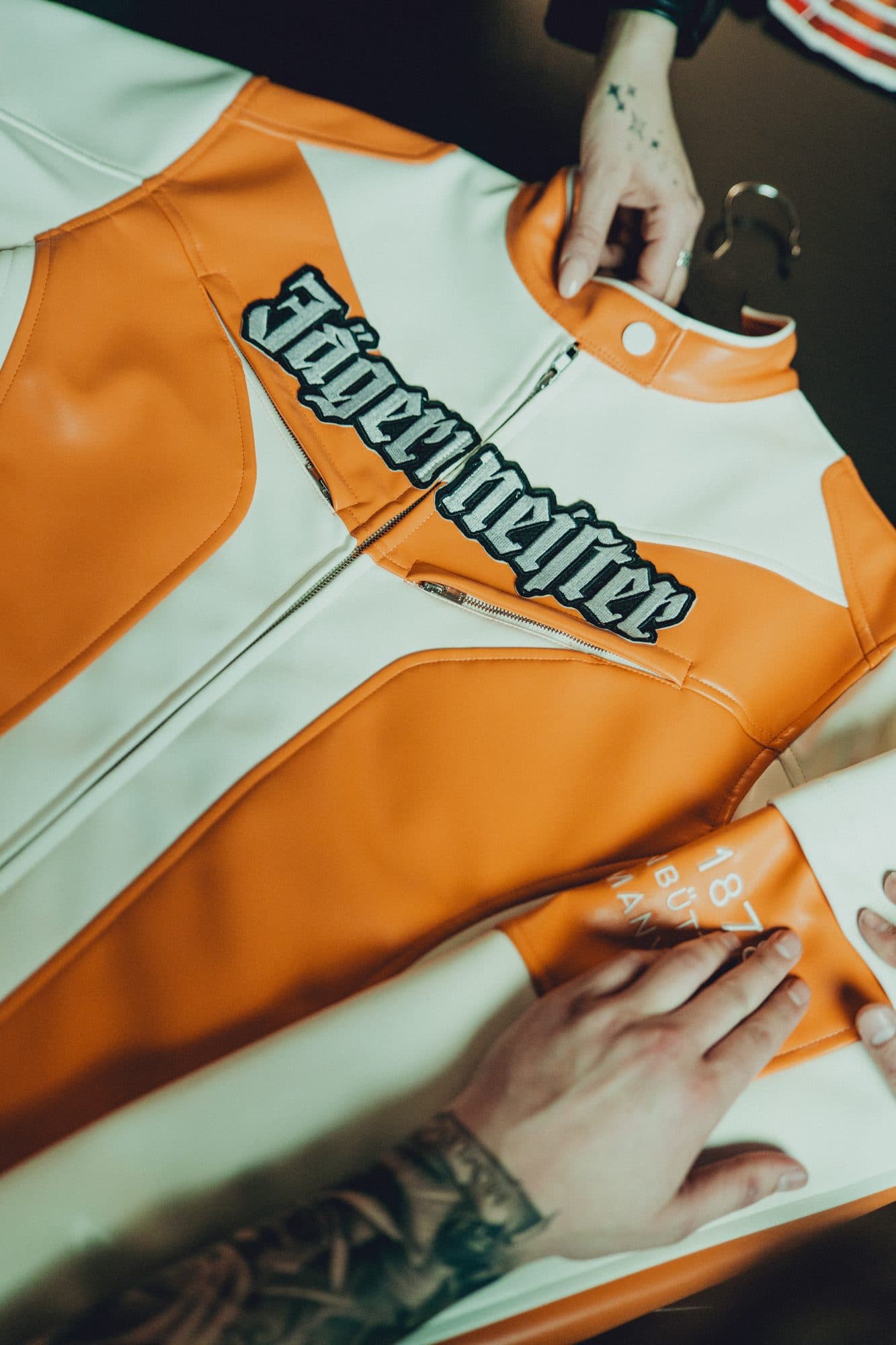 Hands laying out an orange and cream racing jacket with a large embroidered Jägermeister wordmark across the chest.