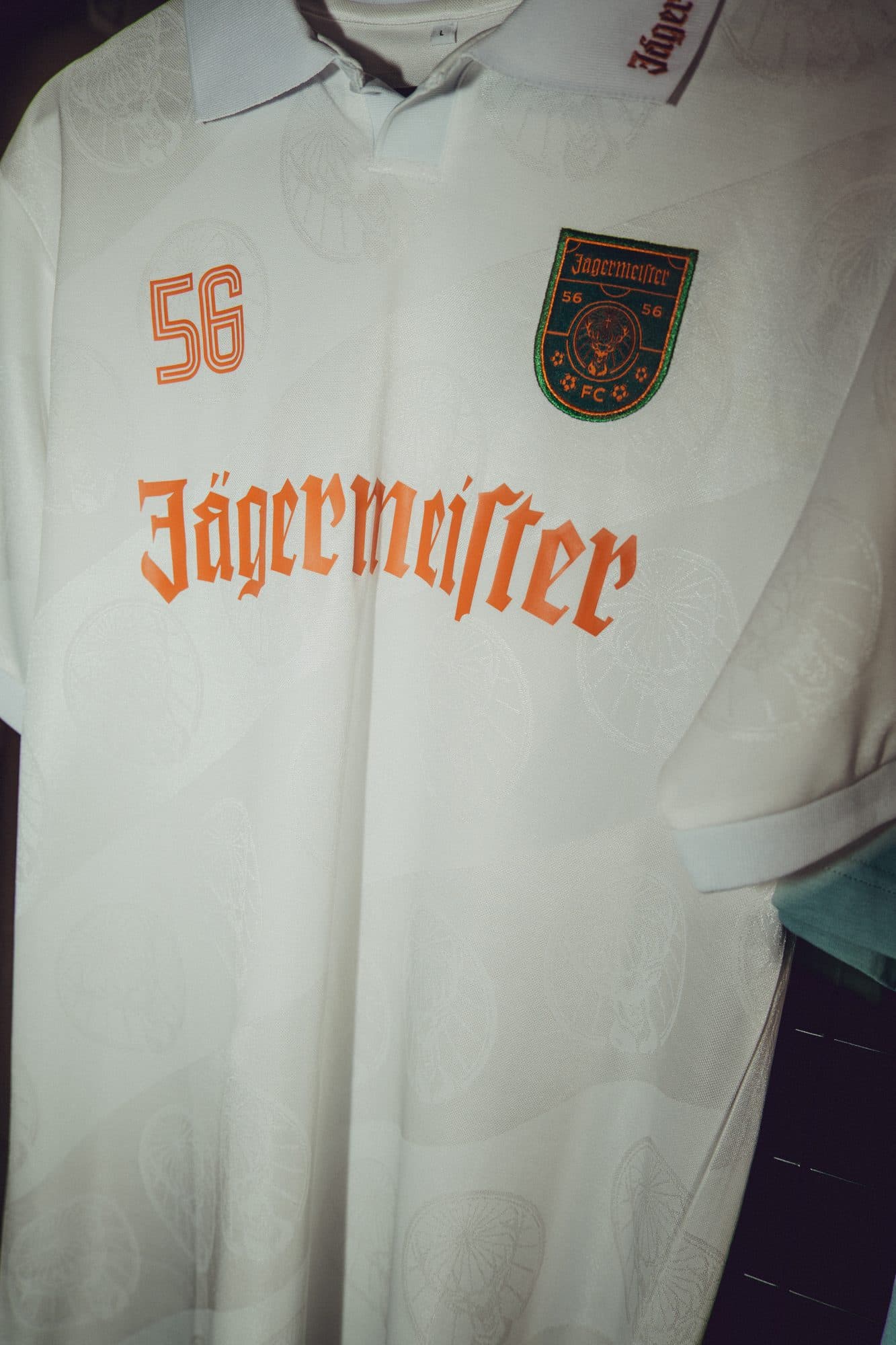 White Jägermeister football shirt with an orange 56 and wordmark, a 56 FC crest badge and a tonal stag roundel pattern in the fabric.