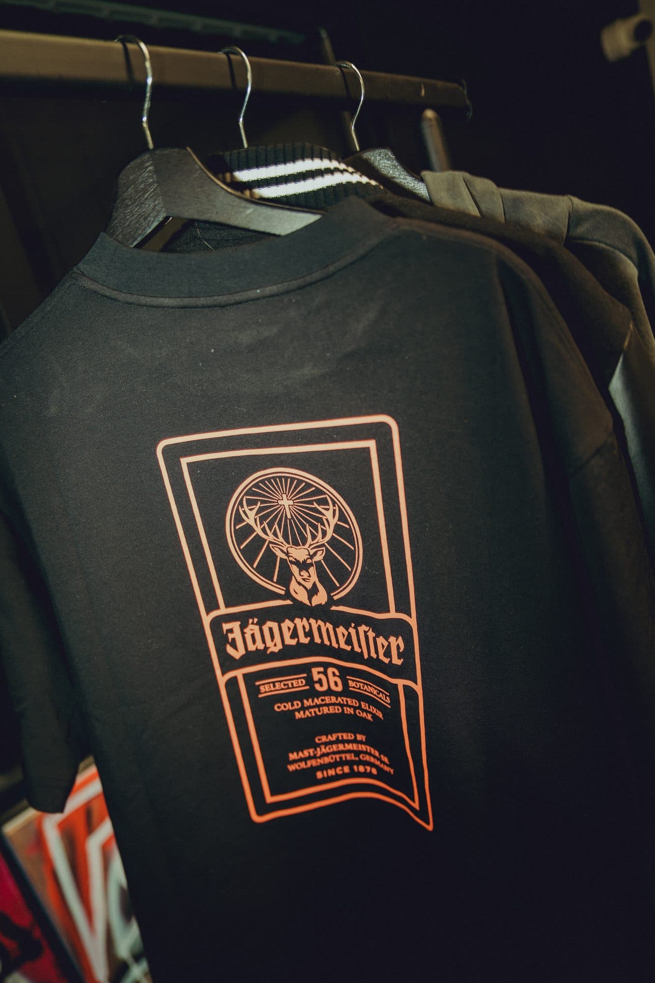 Black tee on a hanger with a large orange Jägermeister back print showing the stag crest above the wordmark and 56 botanicals text.