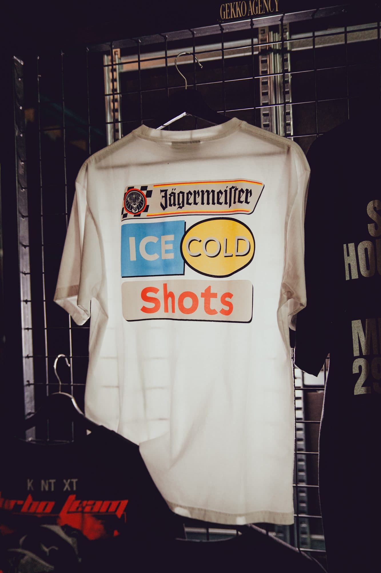 White tee on a hanger against a wire rack, with a Jägermeister Ice Cold Shots racing print on the back.