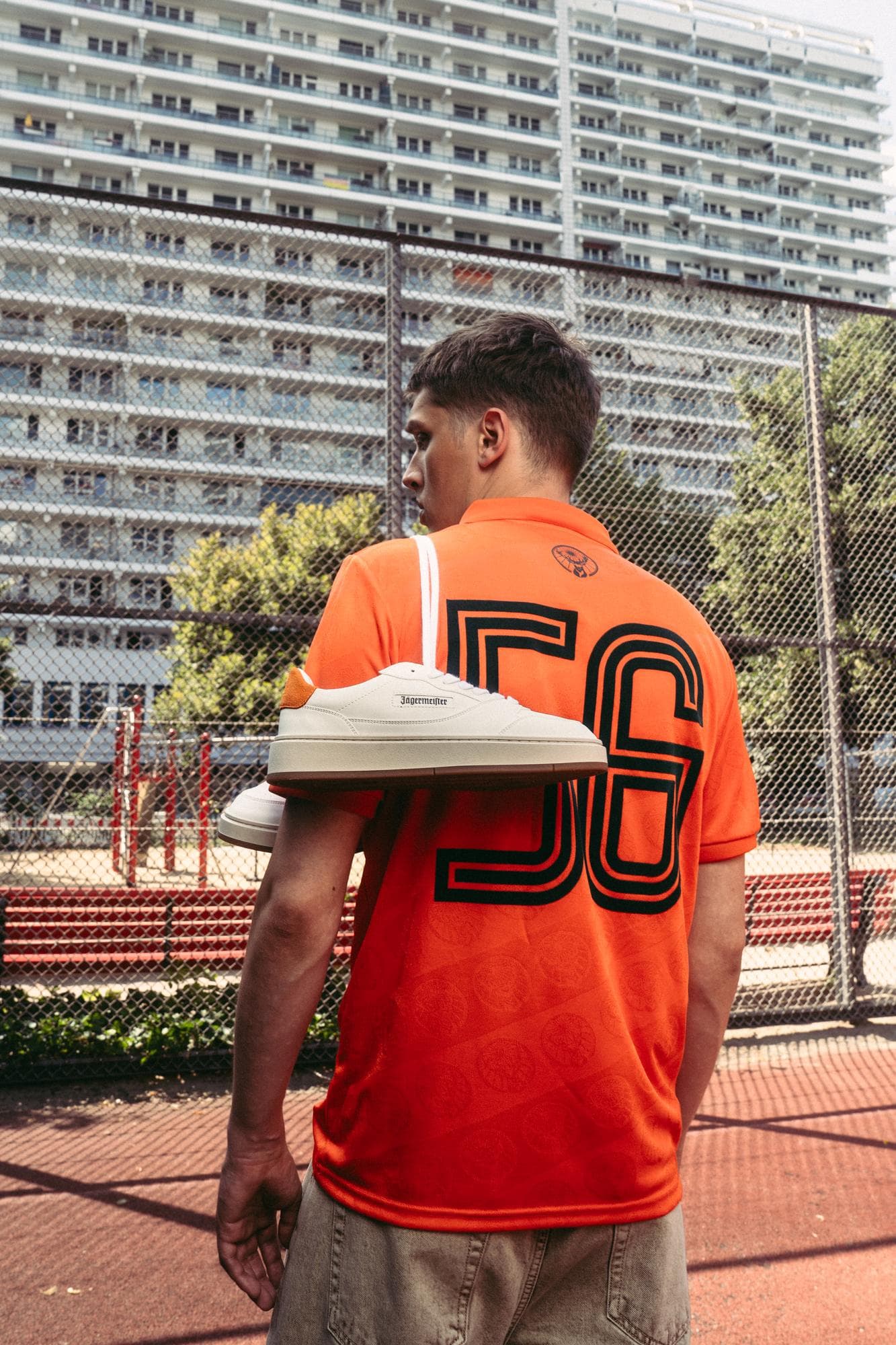 Back of an orange Jägermeister football shirt with a black 56, a white sneaker on the shoulder, on a court between tower blocks.