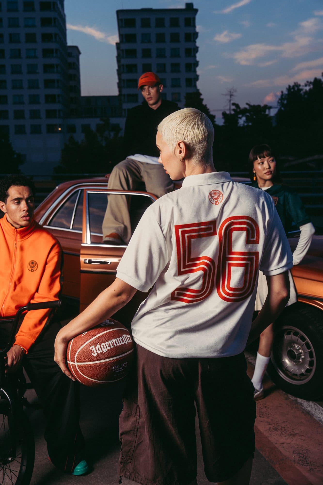 Back of a white Jägermeister shirt with a red 56, a basketball under the arm, beside an orange car.