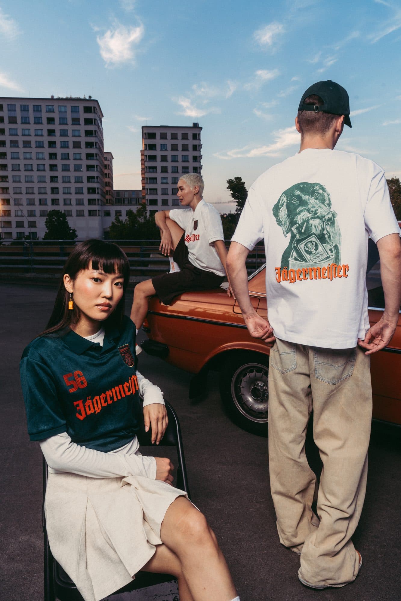 Back of a white T-shirt with a large green stag print and orange Jägermeister lettering, beside a green Jägermeister shirt and an orange car.