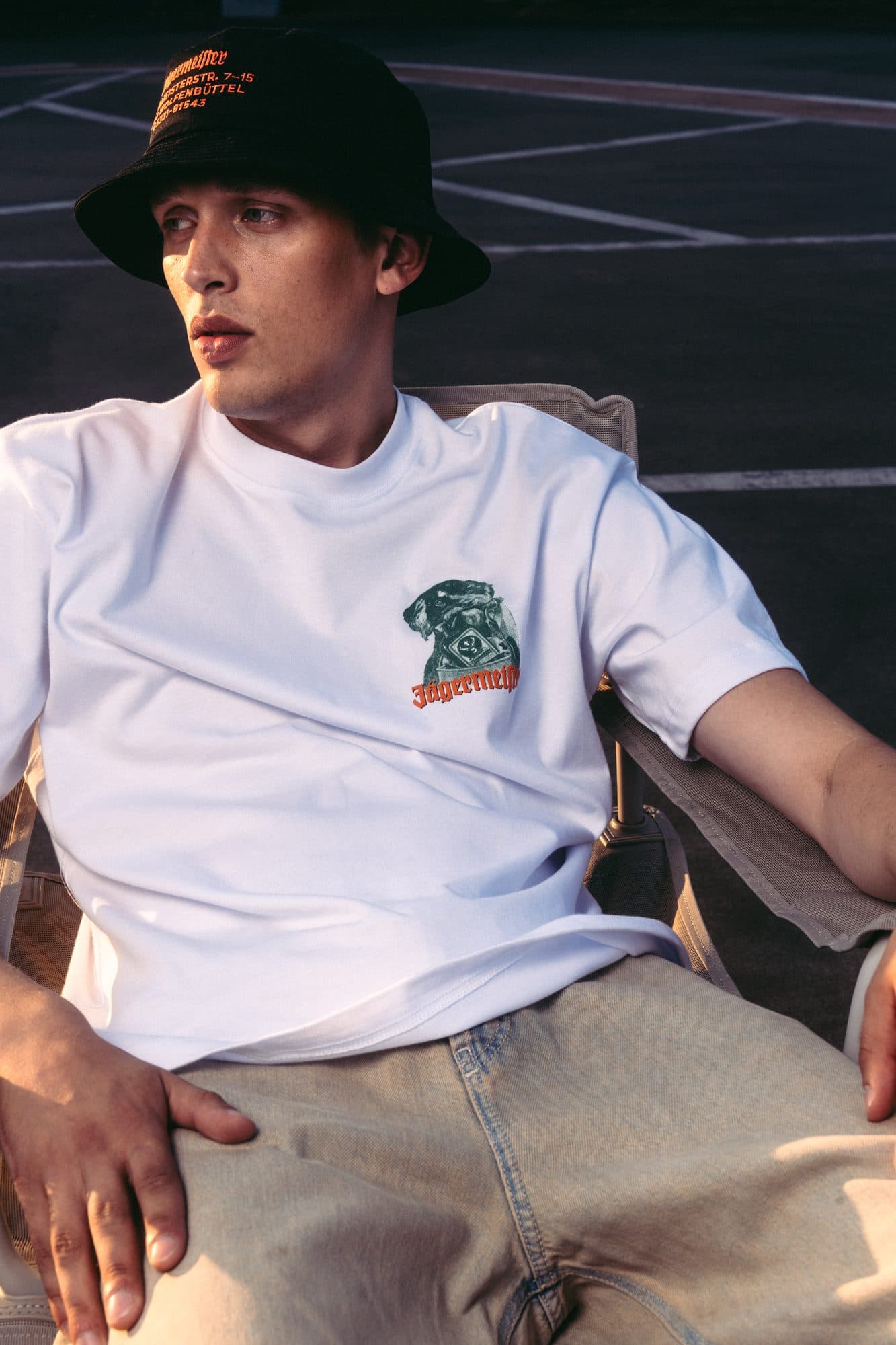 White T-shirt with a green stag-head print on the chest, worn with a black bucket hat.