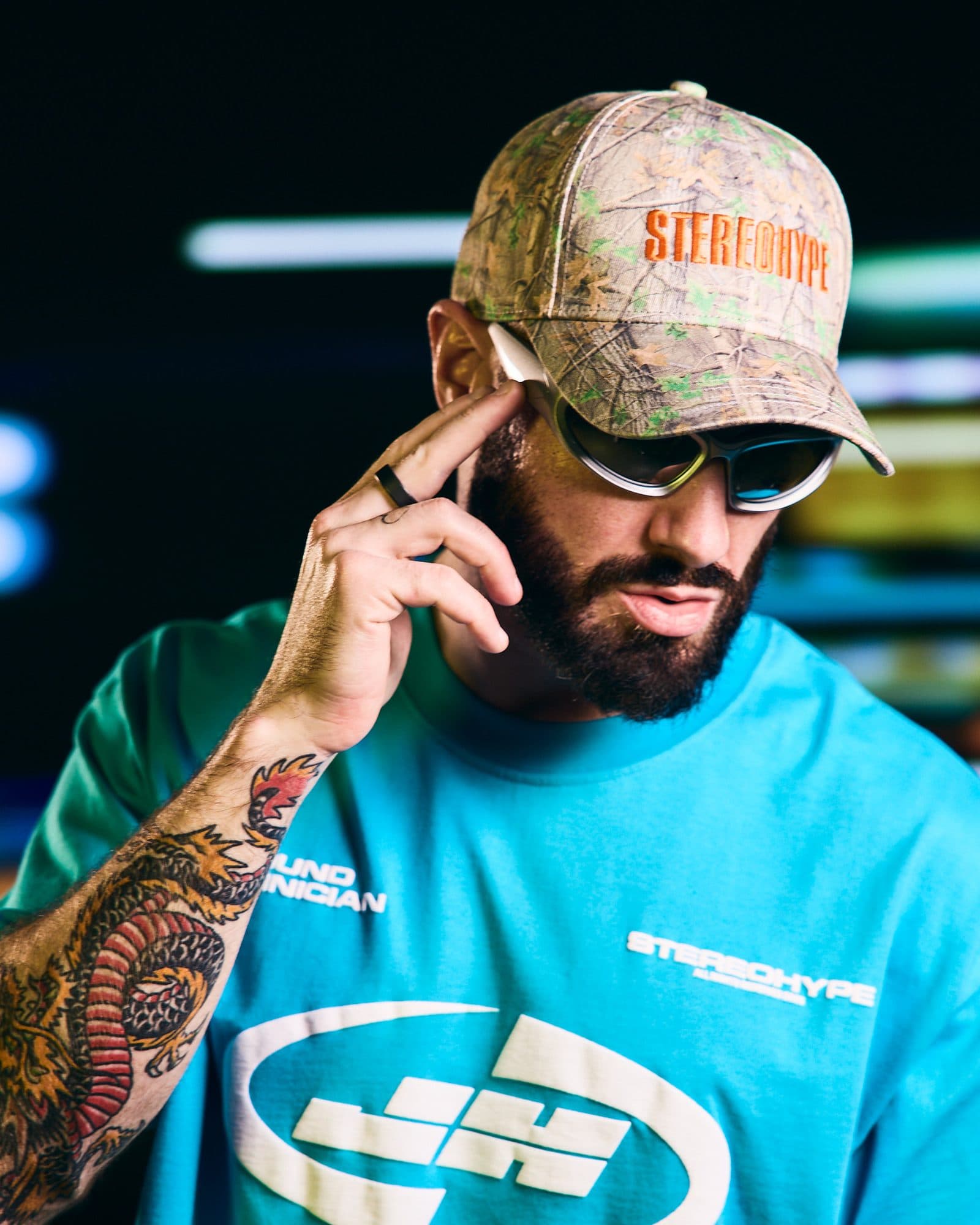 James Hype in sunglasses with one hand at the ear, in a camouflage cap with STEREOHYPE in orange on the front and a turquoise T-shirt with SOUND TECHNICIAN on the chest, partly hidden by his tattooed arm, and STEREOHYPE in small white letters above a large white oval logo, in front of blurred bands of coloured light.