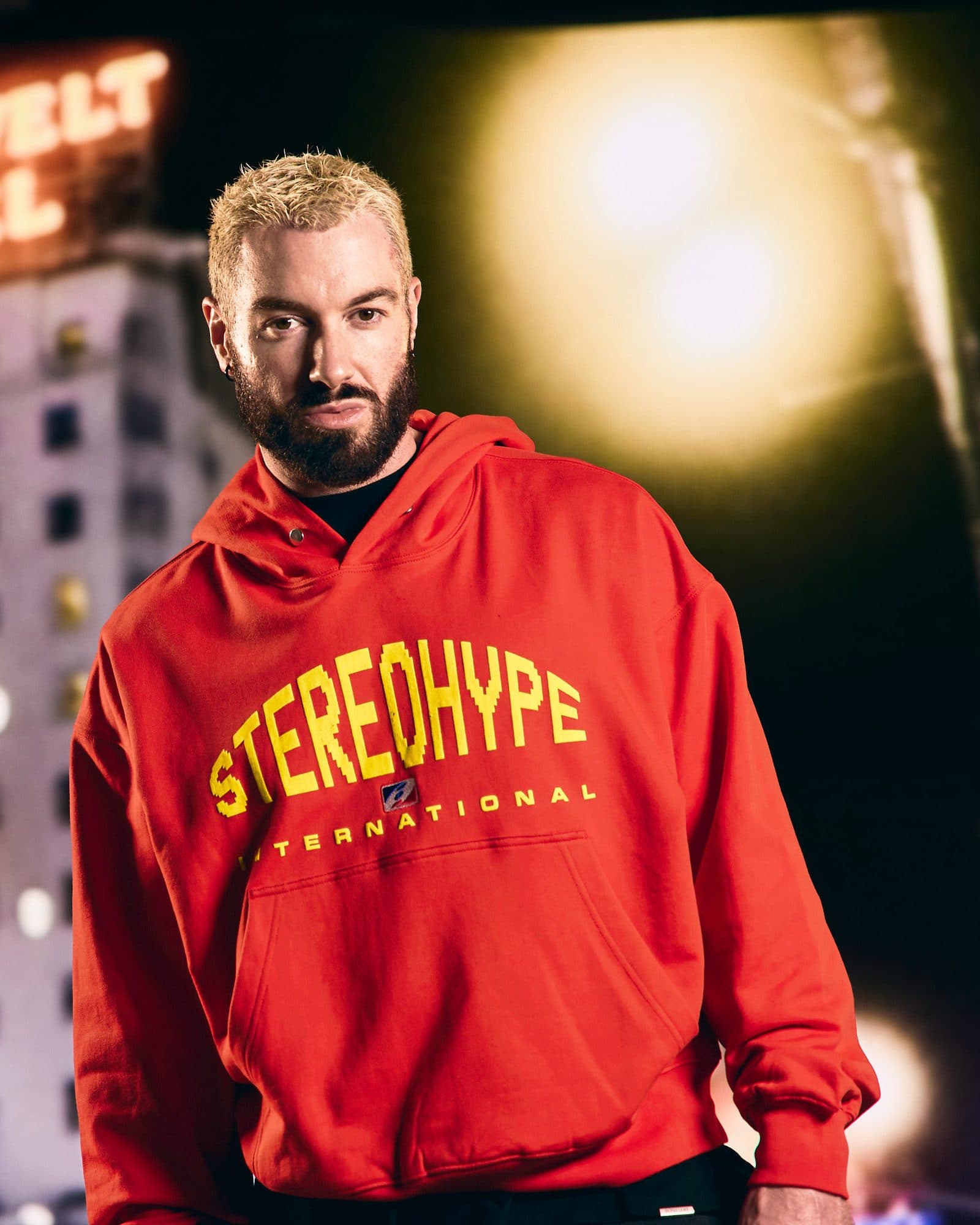 James Hype, with bleached blond hair and a beard, in a red hoodie with STEREOHYPE in large yellow arched letters, a small red, white and blue badge and INTERNATIONAL below, in front of a blurred night scene of a lit high-rise, part of an orange neon sign and a bright light.
