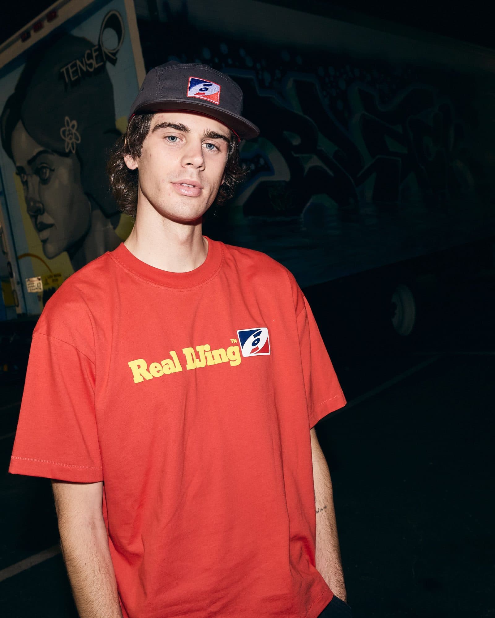 A person in a car park at night in front of a truck painted with a large mural of a face, in a red T-shirt with Real DJing in yellow letters beside a red, white and blue badge and a dark grey cap with the same badge.