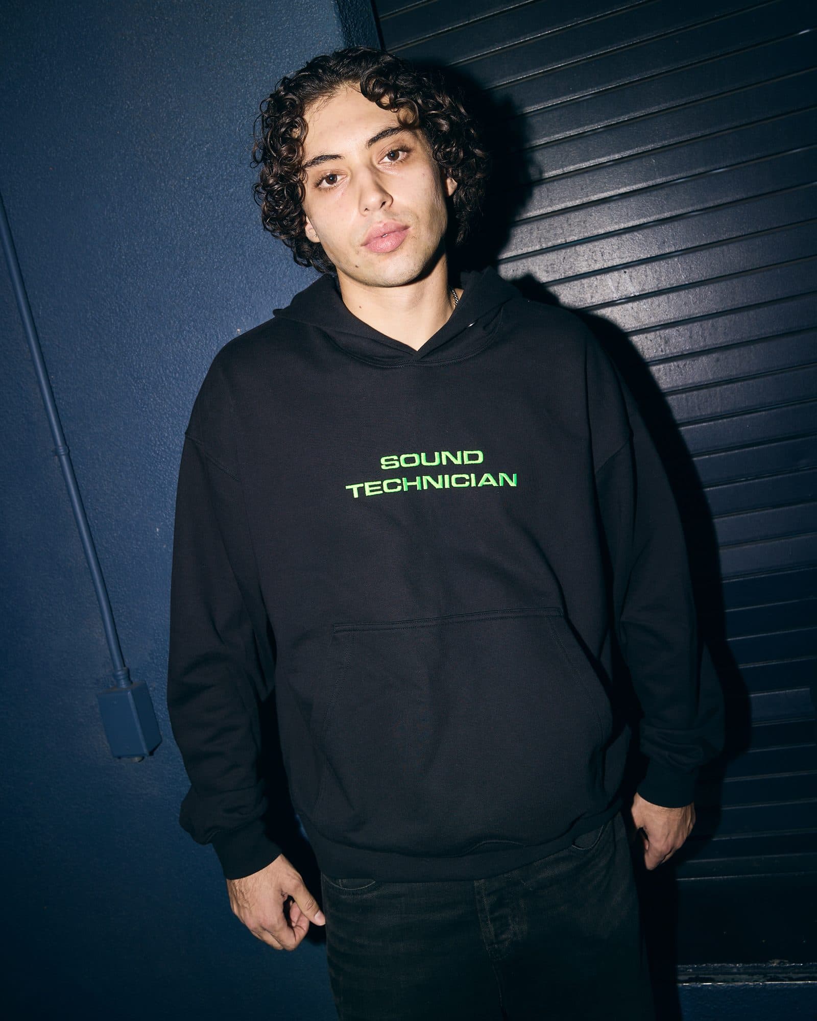 A person with dark curly hair against a dark blue wall and a dark blue roller shutter, in a black hoodie with SOUND TECHNICIAN in green letters on the chest.