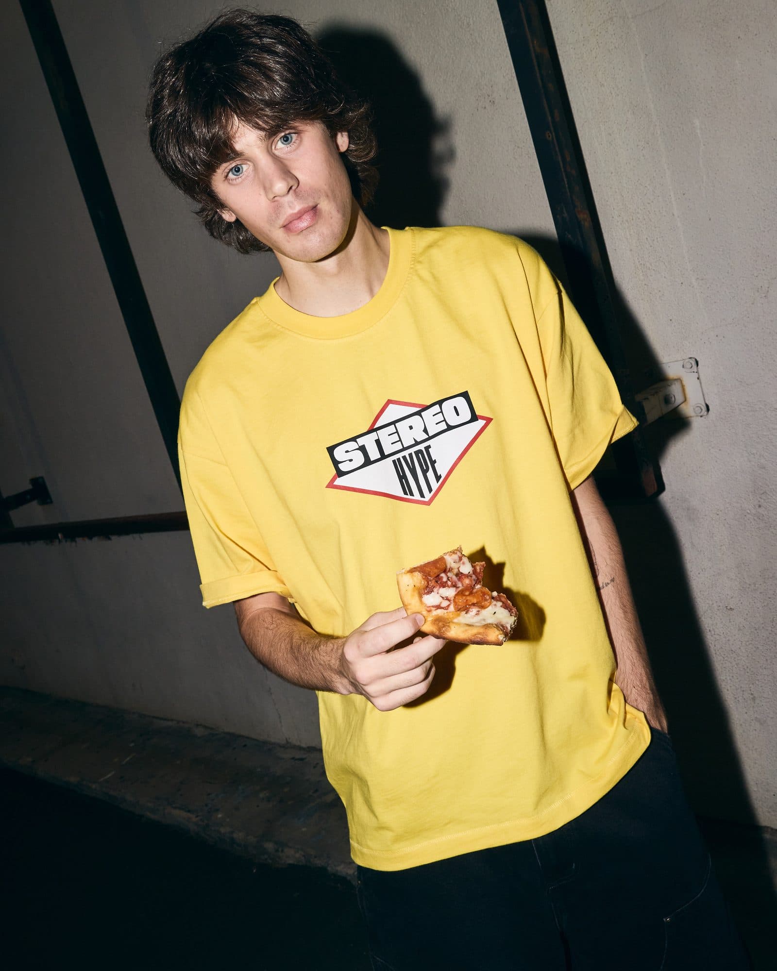 A person lit by a flash against a concrete wall with dark steel beams, holding a slice of pizza, in a yellow T-shirt with STEREO in white on a black band and HYPE in black inside a white diamond edged in red on the chest.