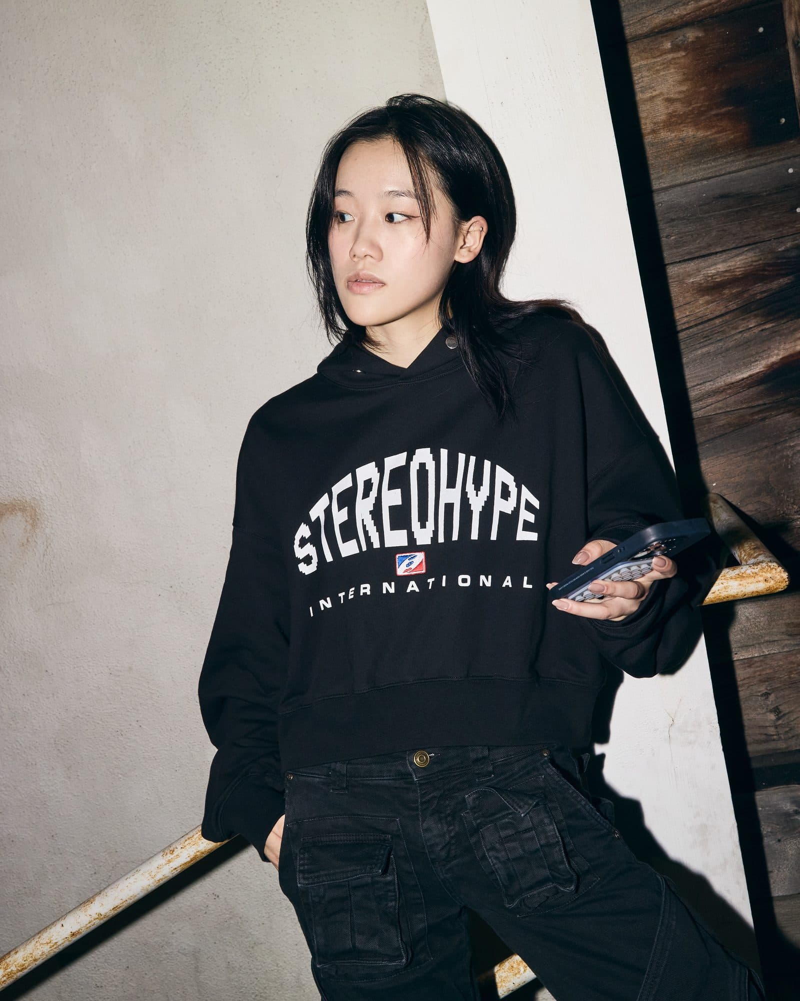 A person holding a phone and leaning on a rusty white handrail by a concrete wall, in a black cropped hoodie with STEREOHYPE in large white arched letters, a small red, white and blue badge and INTERNATIONAL below.