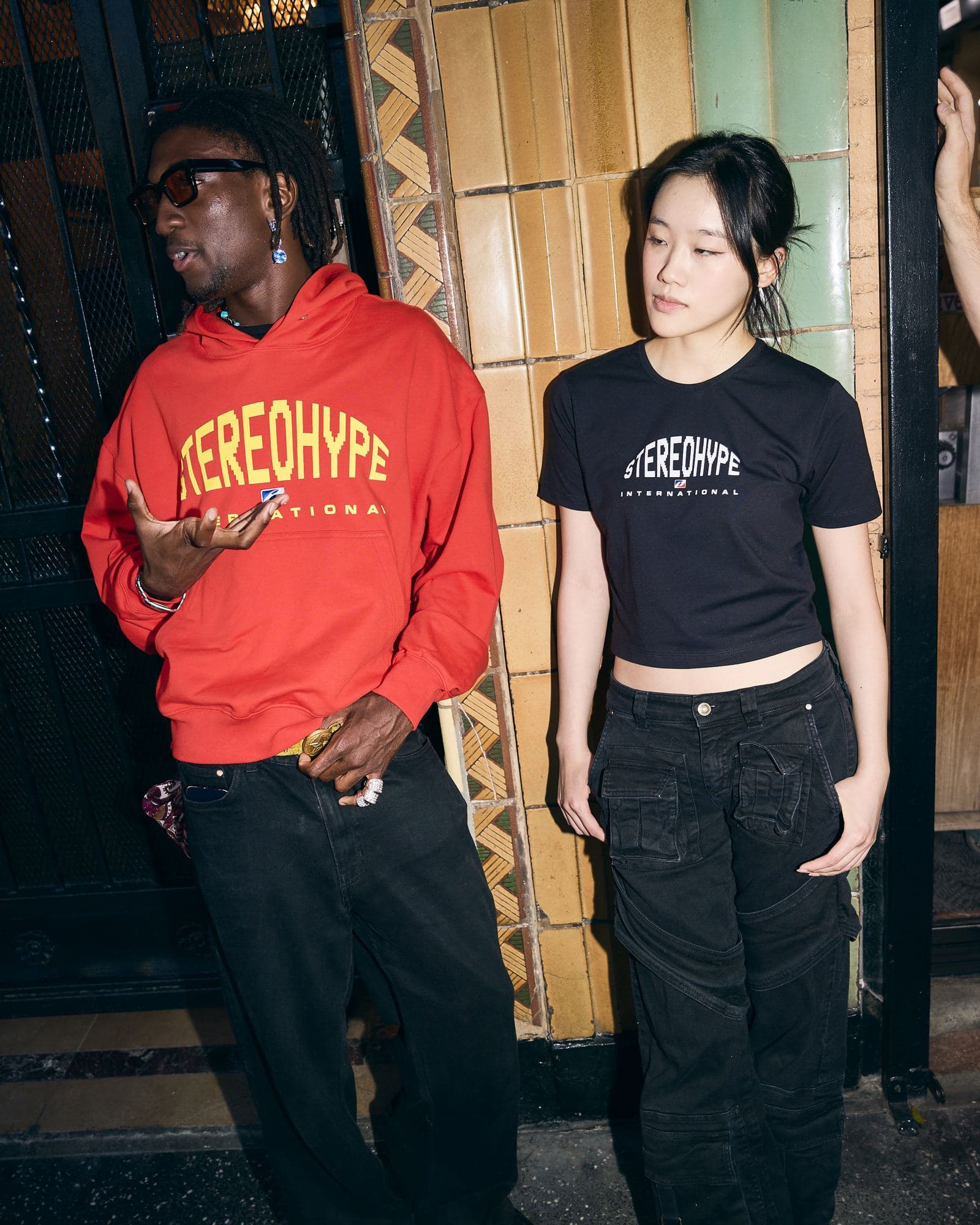 Two people lit by a flash against a wall of yellow and green glazed tiles beside a black metal gate, one in sunglasses and a red hoodie with STEREOHYPE INTERNATIONAL in yellow, one in a black cropped T-shirt with STEREOHYPE INTERNATIONAL in white.