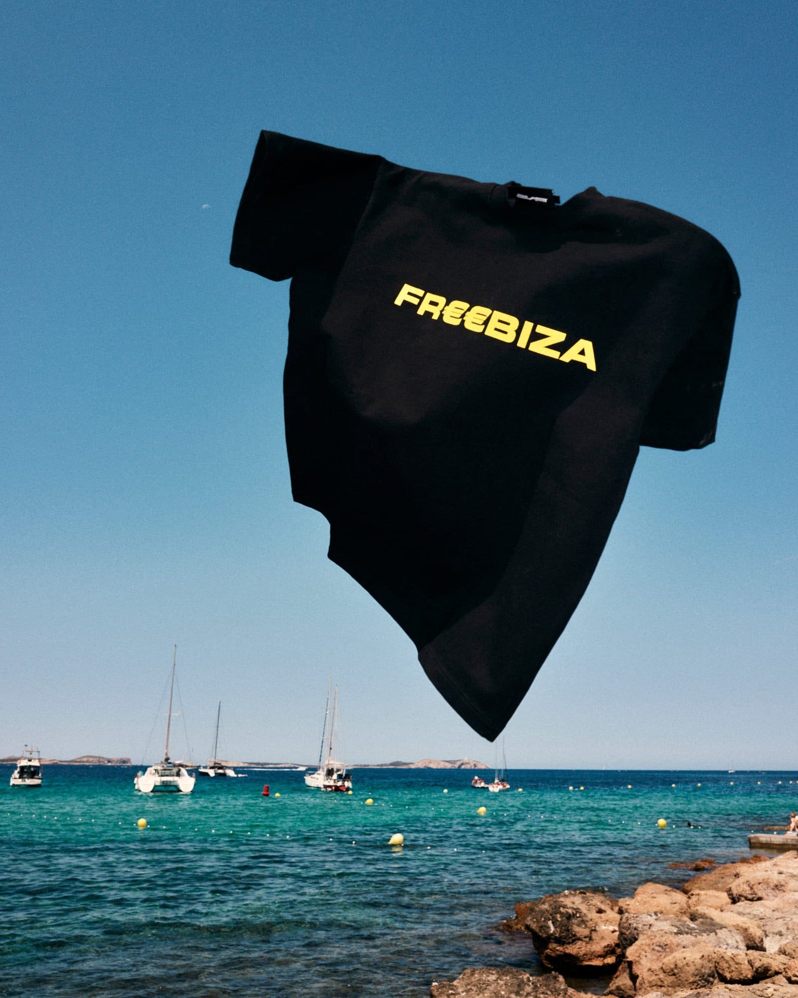 A black T-shirt caught in mid-air against a clear blue sky, printed in yellow across the chest with FREEBIZA, its two E's drawn as euro signs, above a turquoise sea with sailboats and yellow buoys and brown rocks in the foreground.