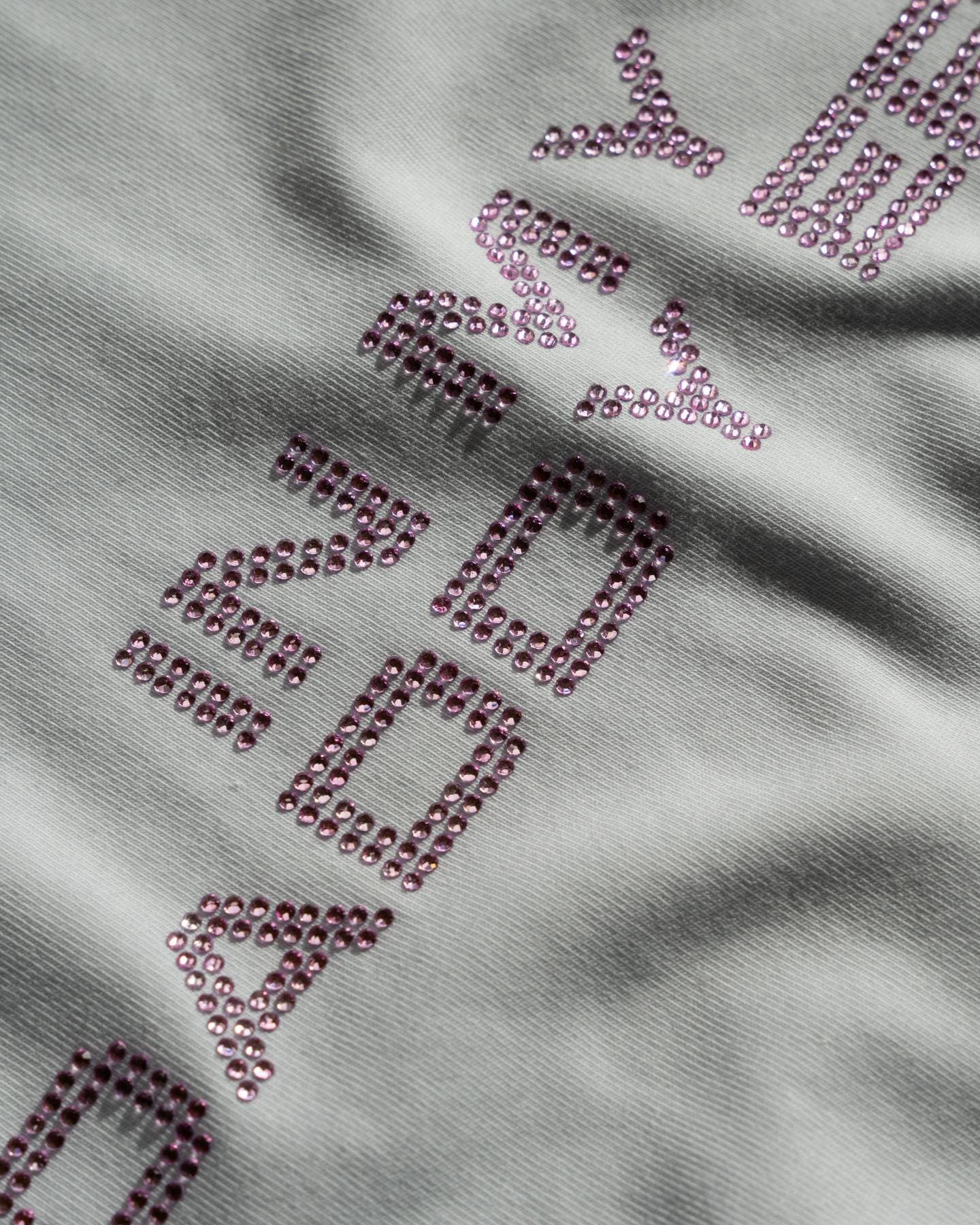 Close-up of IN MY DADDY ERA in pink rhinestones, set at an angle on creased pale grey T-shirt fabric in hard sunlight, the ends of the lettering cut off by the frame.