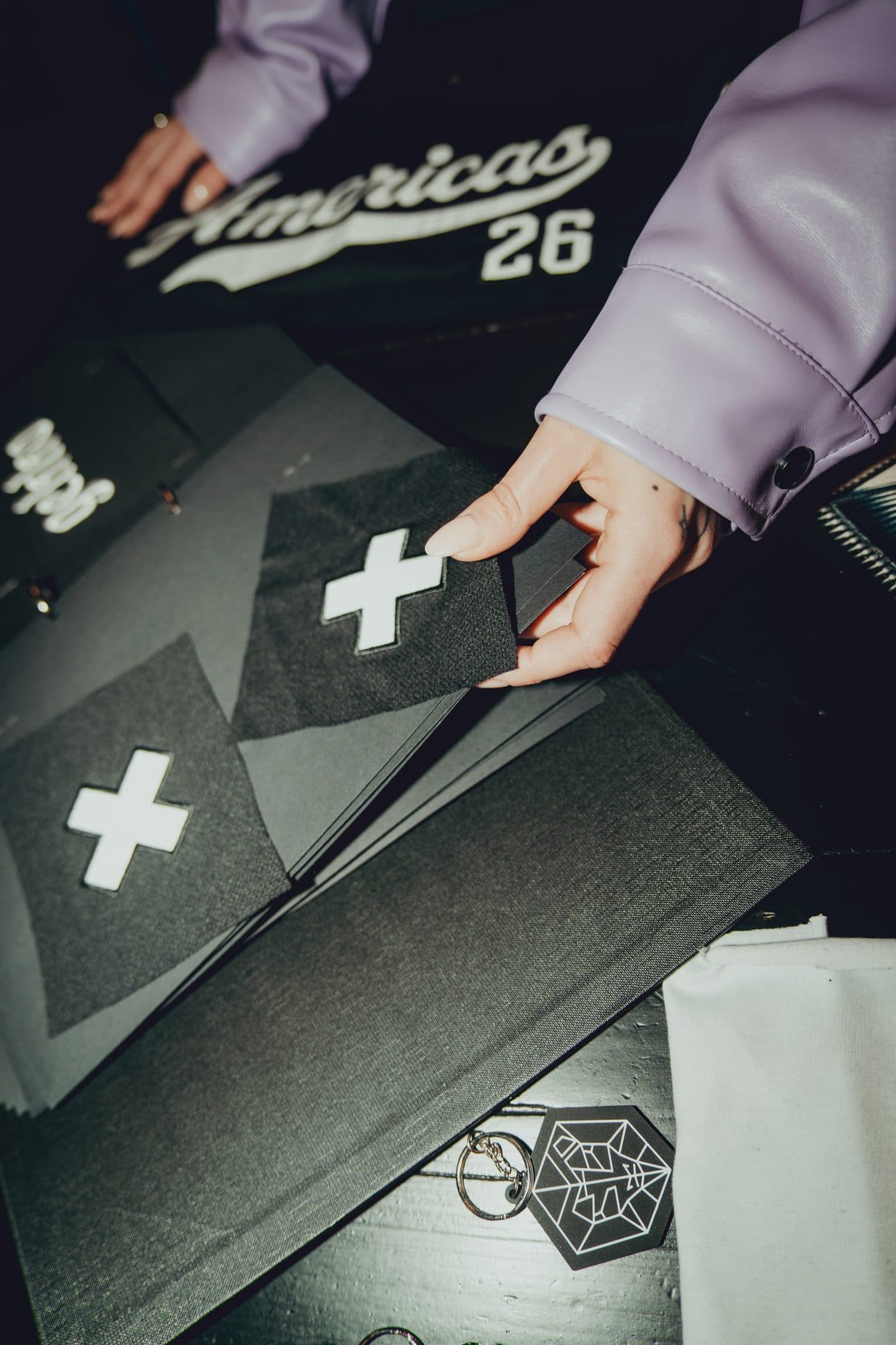 Hand in a lilac leather sleeve holding a black fabric swatch with a white cross patch over a sample binder, with an Americas 26 jersey and a hexagon keychain on the table.