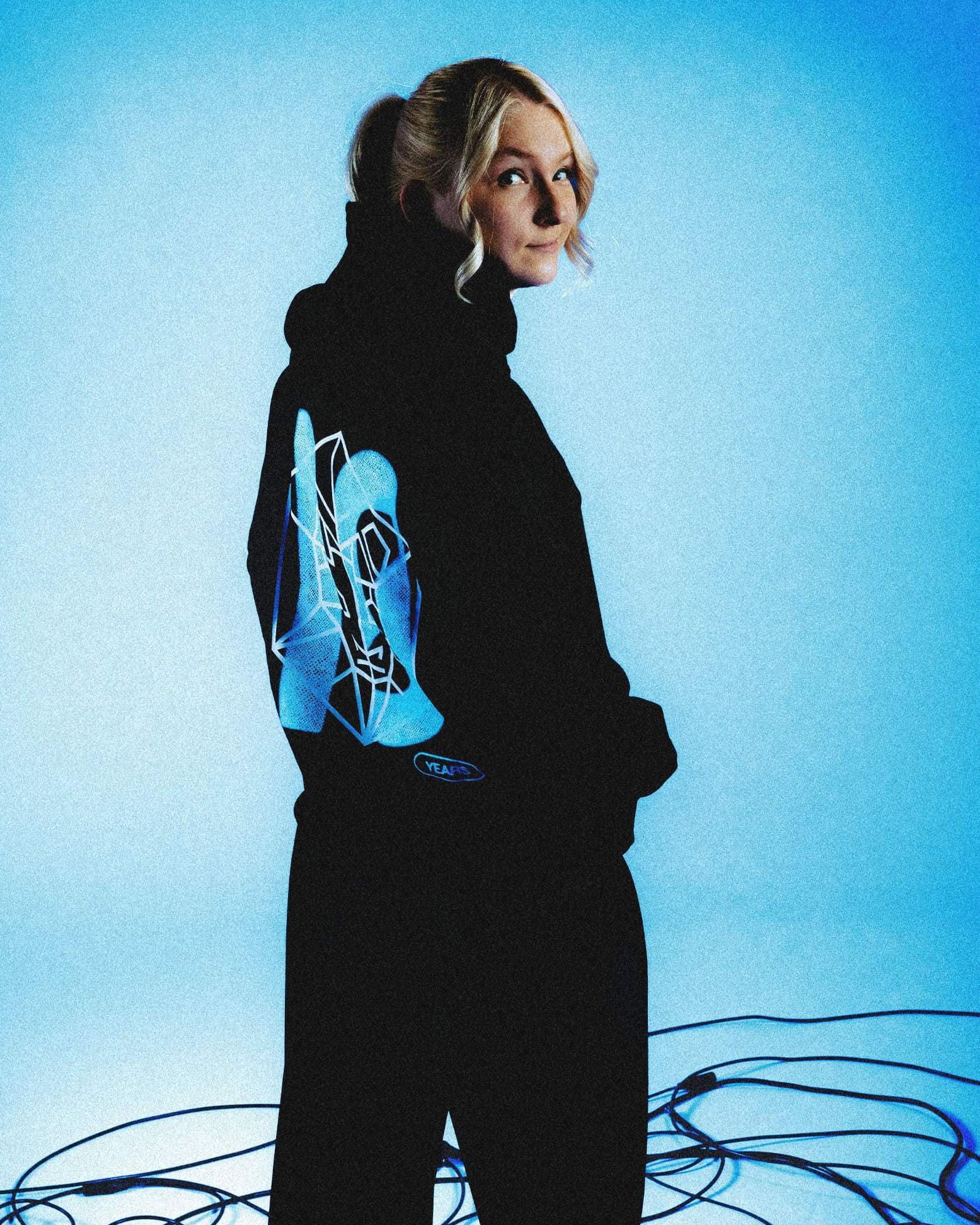 A person looking back over their shoulder in a blue-lit studio among black cables, in a black hoodie with a large blue 10 and a white wireframe emblem printed on the back and a small YEARS badge.