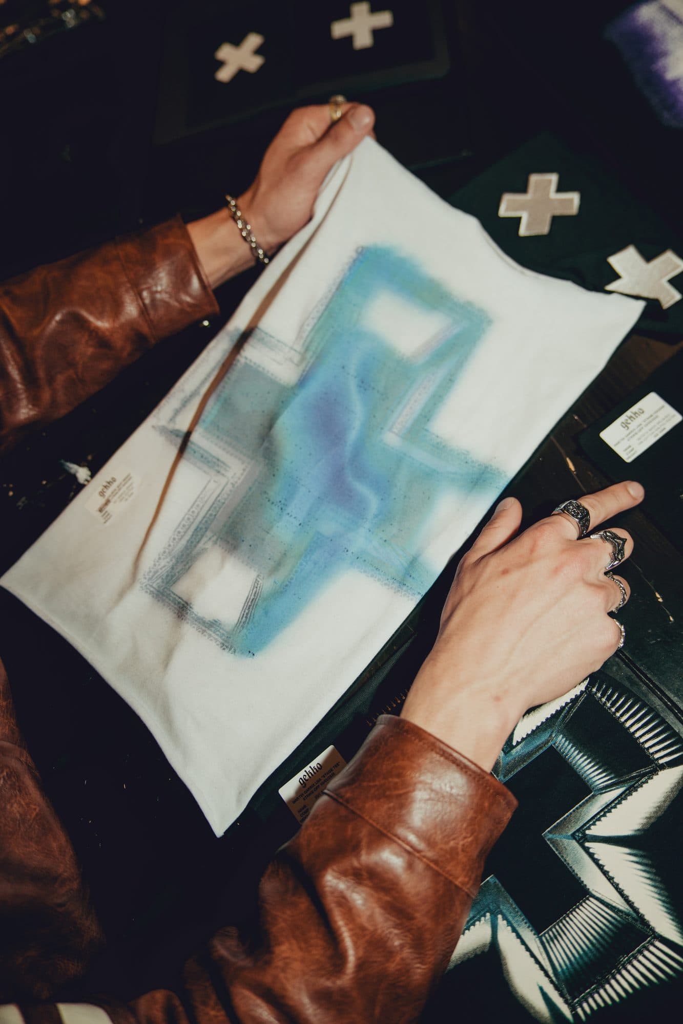 Hands in a brown leather jacket holding a white tee panel with a blue airbrushed cross print, with white embroidered crosses on black fabric around it.
