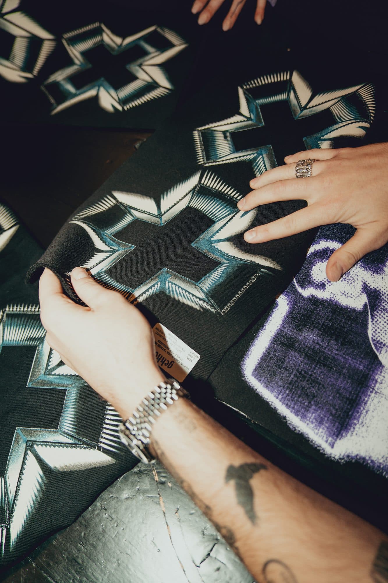 Hands with a steel watch and a silver ring holding a black test panel with a chrome-effect cross print, beside a purple halftone print.