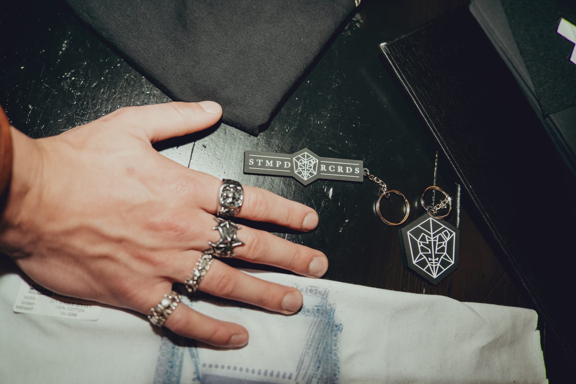 Ringed hand on a dark table next to two STMPD RCRDS rubber keychains, one a bar and one a hexagon, with a white printed sample below.