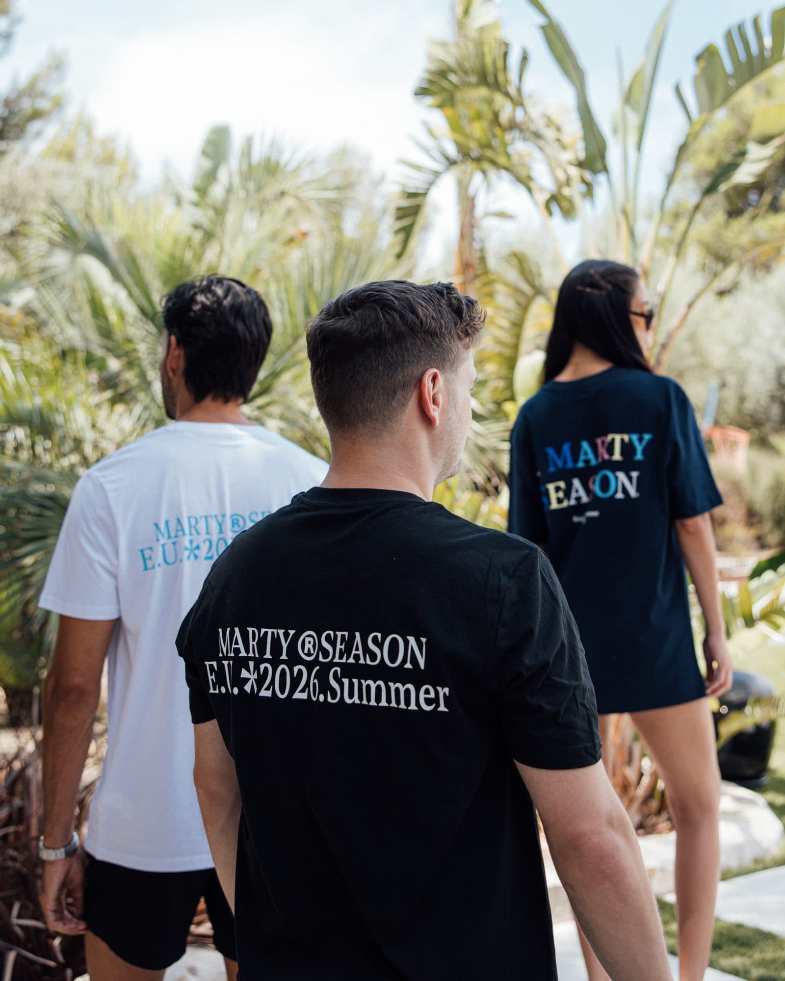 Three people seen from behind among palms, in a white T-shirt with a light blue MARTY SEASON print, a black T-shirt with MARTY SEASON E.U. 2026 Summer printed across the back in white and a navy T-shirt with MARTY SEASON in coloured letters.