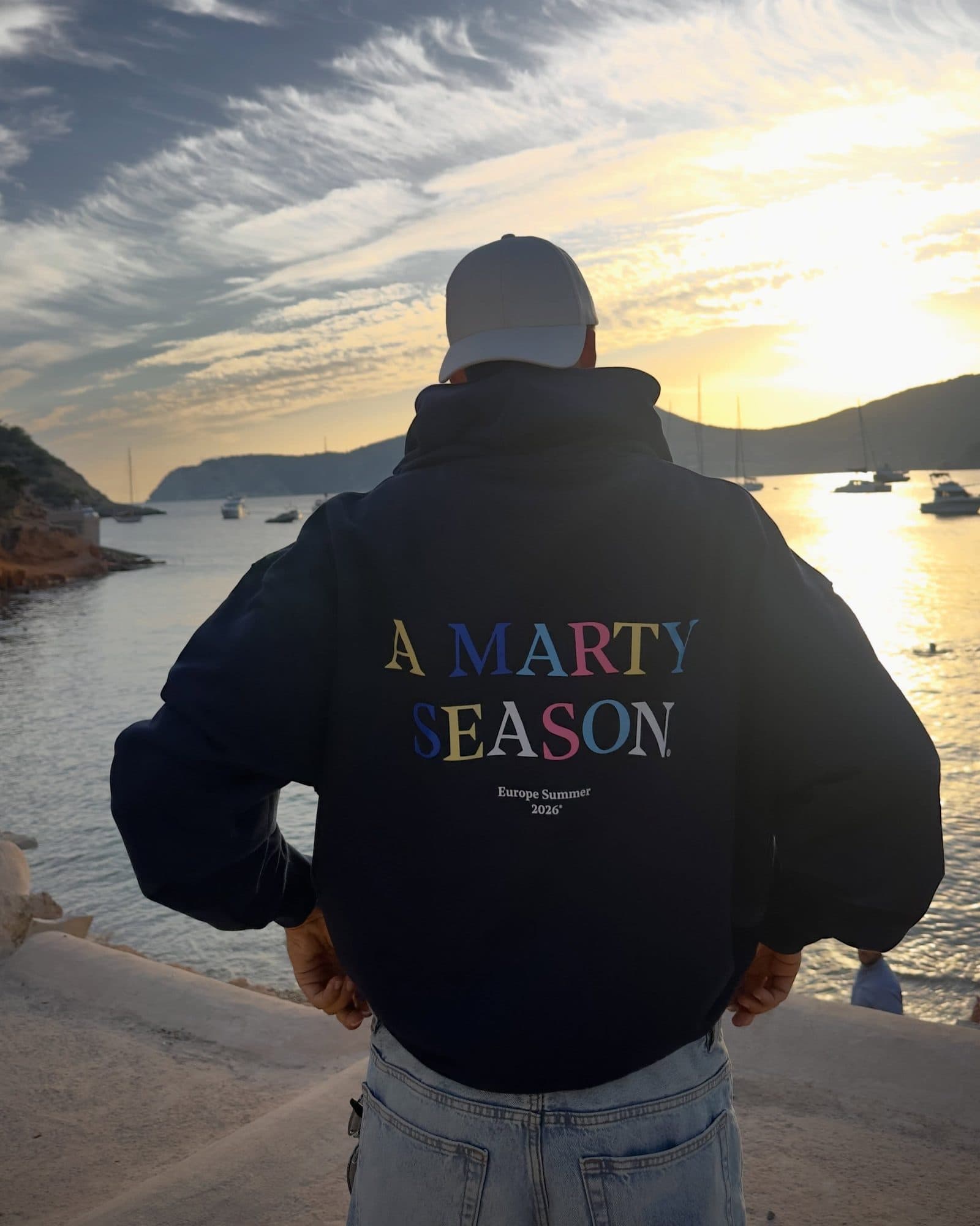 A person in a cap seen from behind above a bay at sunset, hands on hips, in a navy hoodie with A MARTY SEASON in coloured letters and Europe Summer 2026 printed on the back.