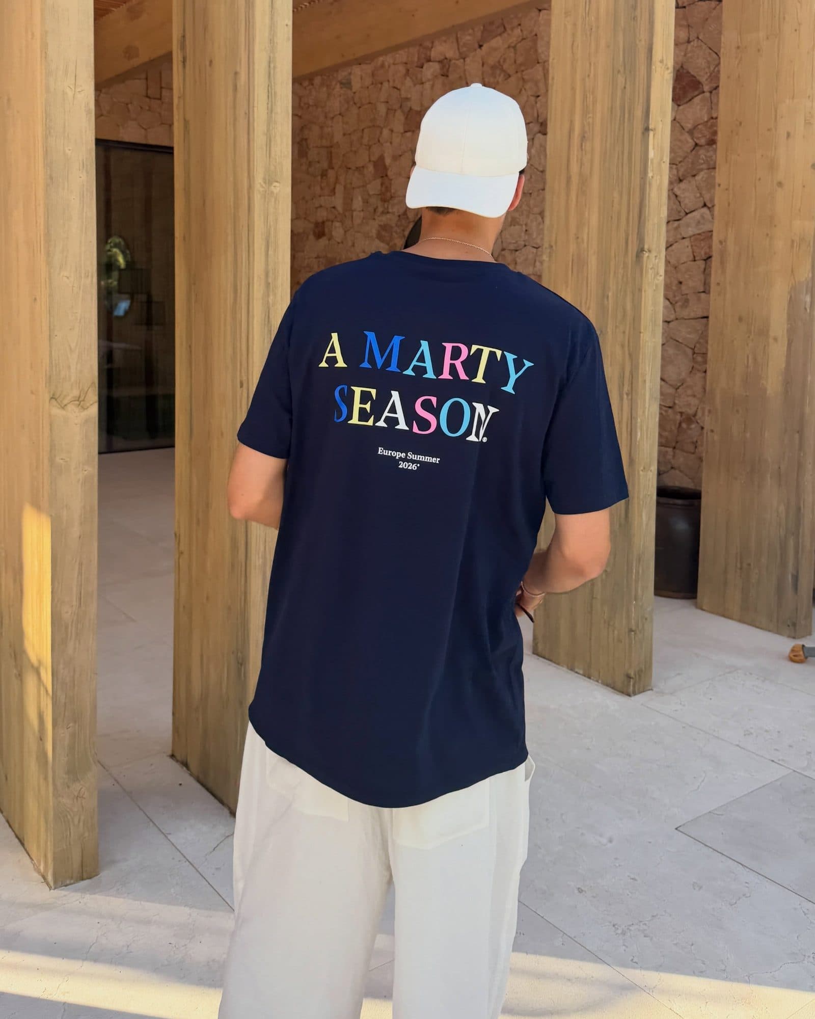 A person in a white cap seen from behind under a wooden pergola, in a navy T-shirt with A MARTY SEASON in coloured letters and Europe Summer 2026 printed on the back.