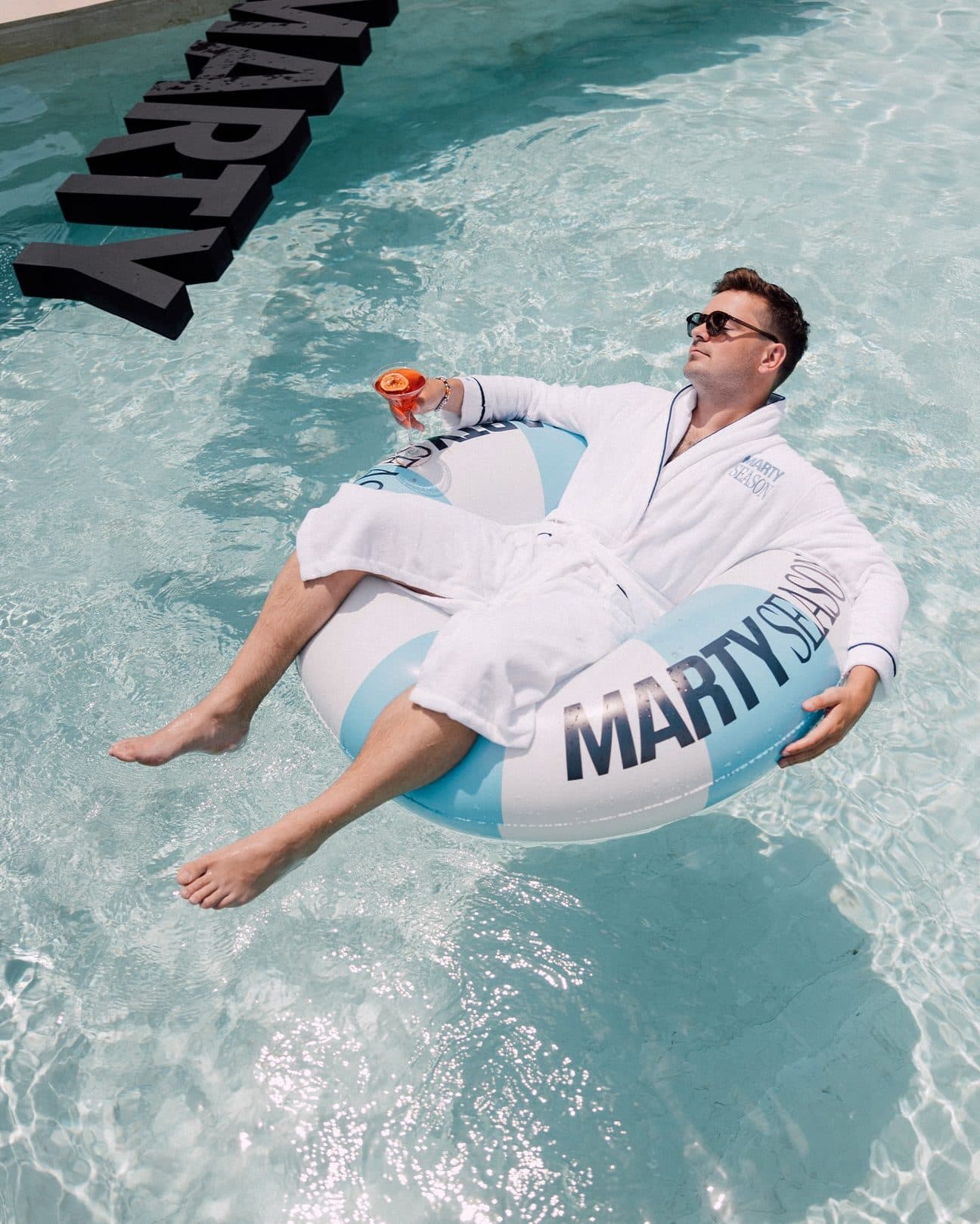 A person in sunglasses and a white MARTY SEASON bathrobe lying back on a light blue and white MARTY SEASON swim ring in a pool, a drink in hand, large black MARTY letters floating beside it.