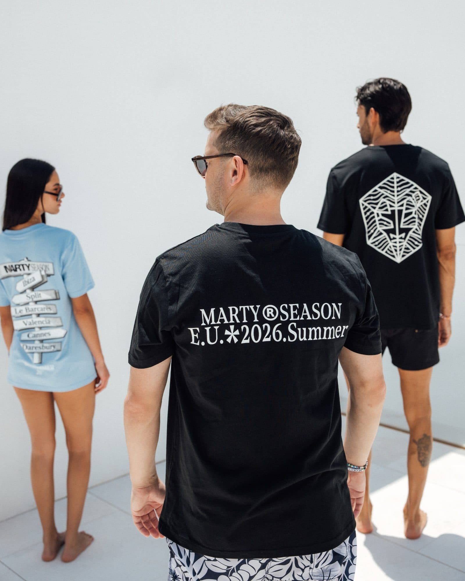 Three people seen from behind on a white terrace, in a black T-shirt with MARTY SEASON E.U. 2026 Summer printed across the back, a light blue T-shirt with a signpost print of summer destinations and a black T-shirt with a large white hexagonal emblem.