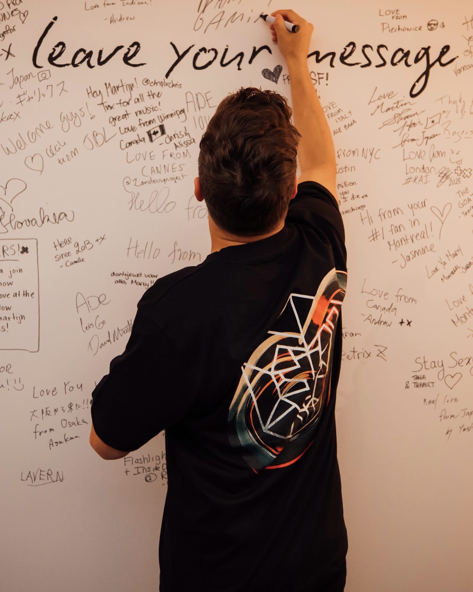 A person seen from behind writing on a wall of handwritten fan messages under the words leave your message, in a black T-shirt with a large white wireframe emblem over orange and dark swirls on the back.