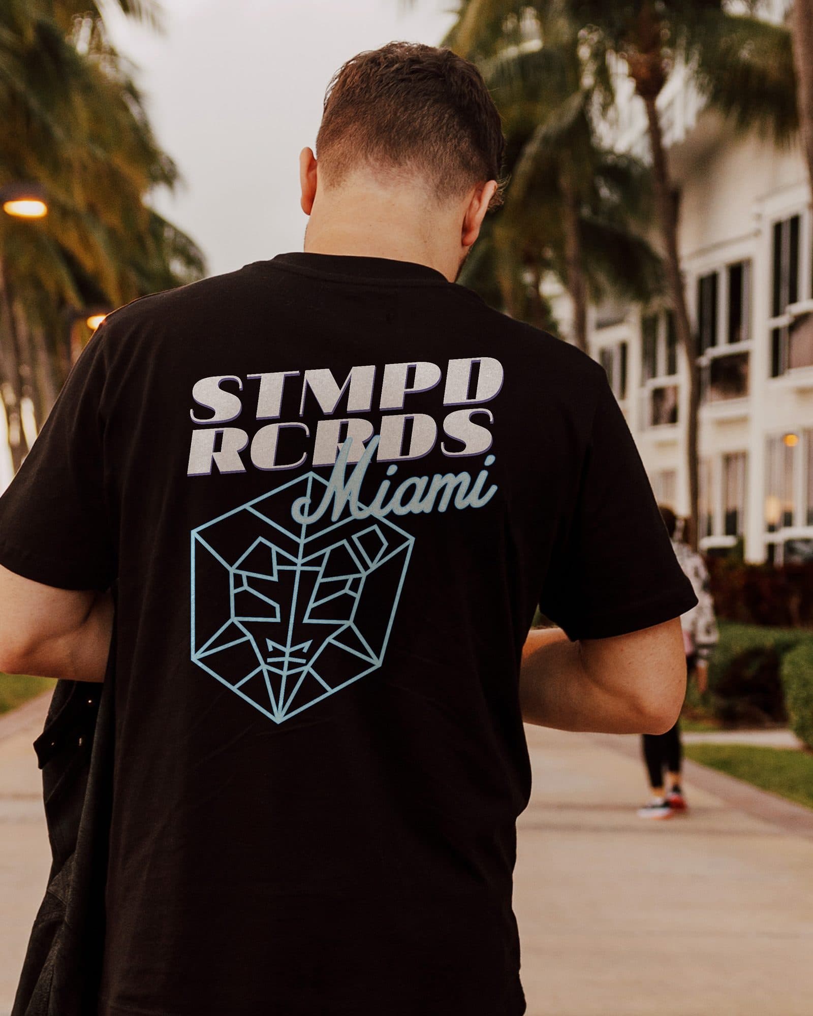 A person seen from behind on a path lined with palm trees, in a black T-shirt with a STMPD RCRDS Miami back print and the label's emblem.