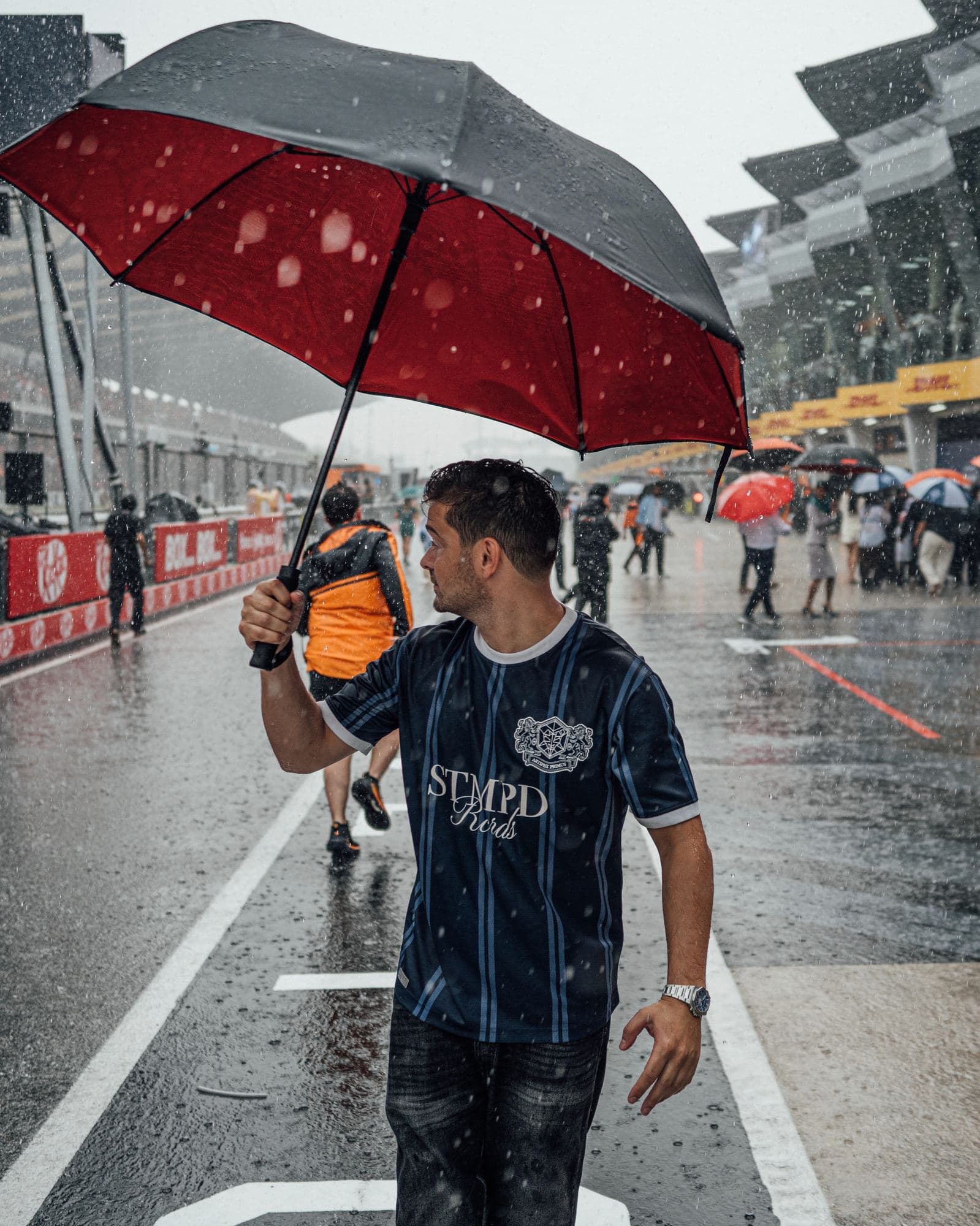 A person under a red and grey umbrella in a rain-soaked pit lane, in a navy pinstripe jersey with STMPD Rcrds lettering and a crest of two lions around the label's emblem.