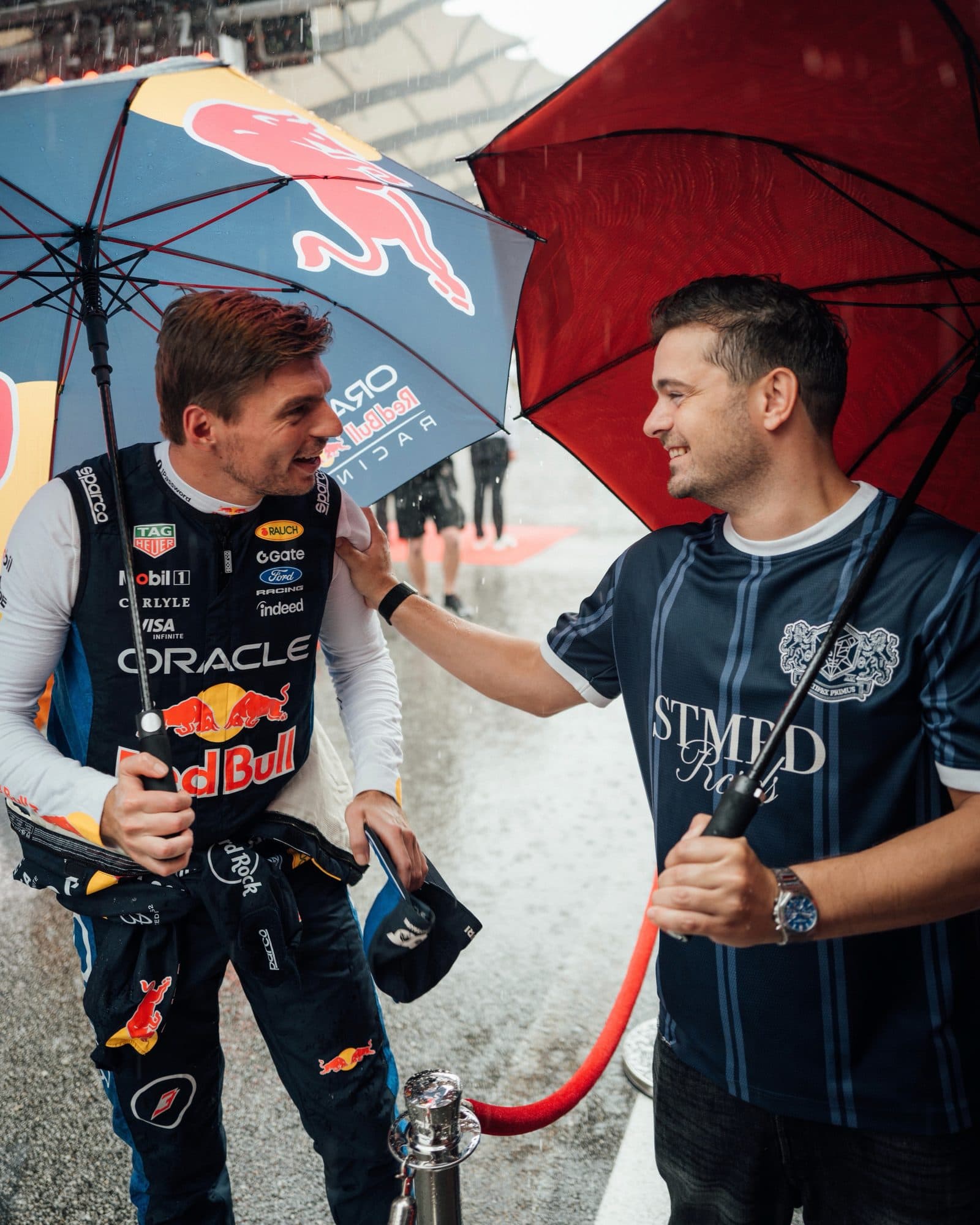 Two people laughing under umbrellas in the rain: one in a racing suit covered in sponsor logos, the other in a navy pinstripe STMPD Rcrds jersey with a crest, a hand on the first one's shoulder.