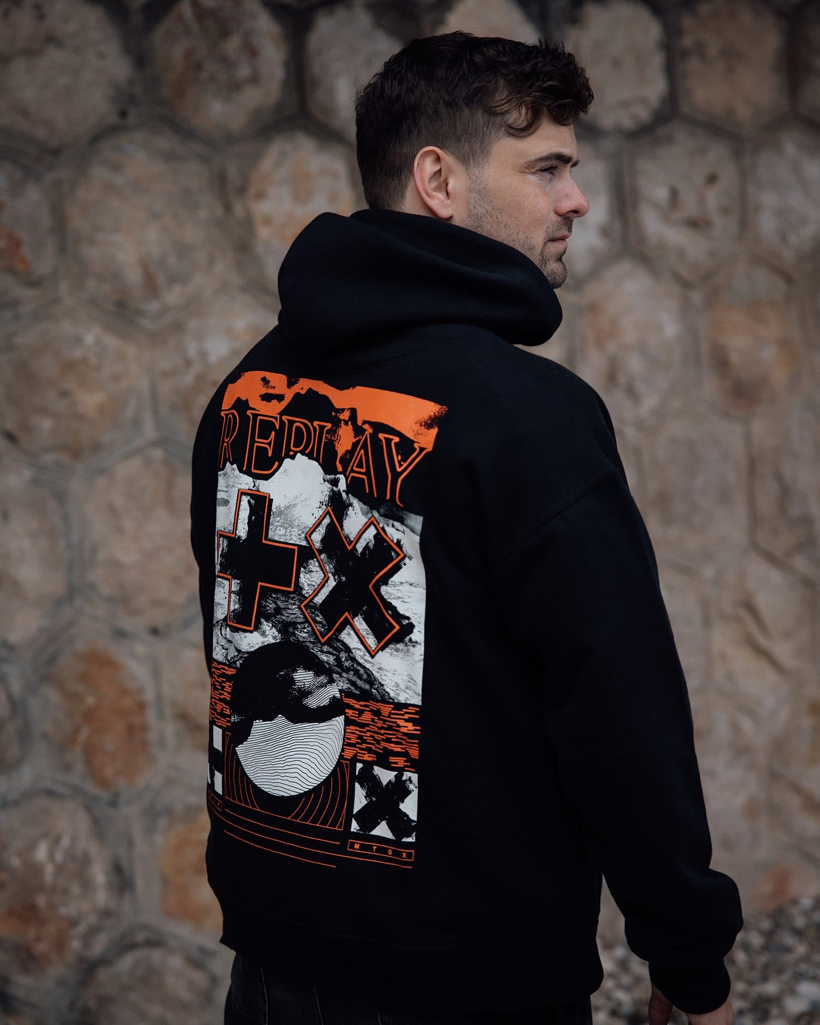 Back of a black hoodie with a large orange, black and white Replay print, a plus and an X over a rocky landscape, worn by a person in profile in front of a stone wall.
