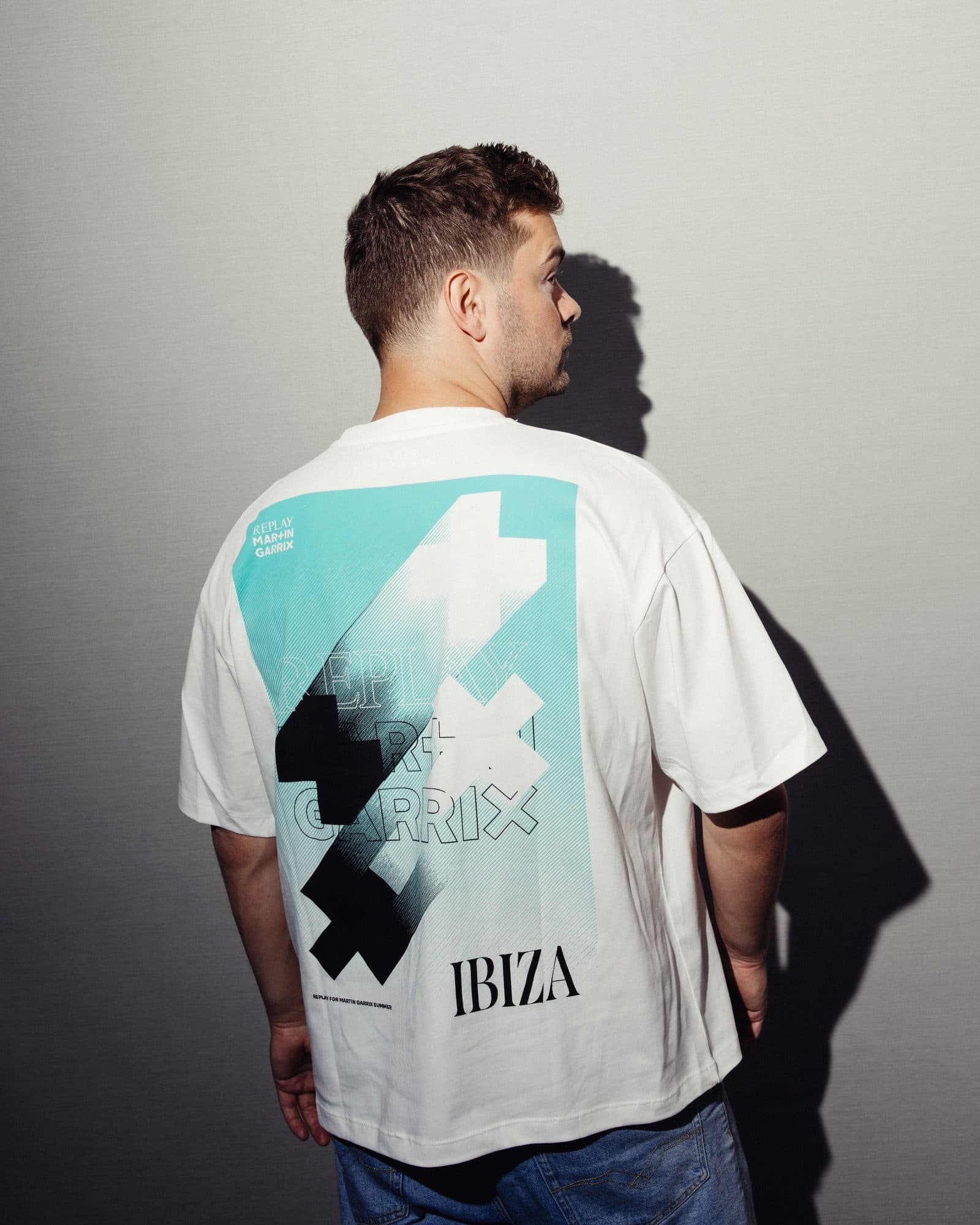 Back of a white T-shirt with a large turquoise, black and white print of plus and X shapes, outlined REPLAY MARTIN GARRIX lettering and IBIZA, worn by a person in profile against a grey wall.