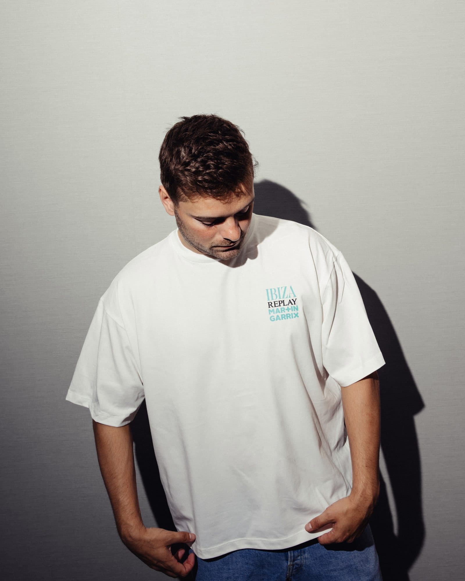 A person looking down against a grey wall, in a white T-shirt with a small IBIZA REPLAY MARTIN GARRIX print in turquoise and black on the chest.