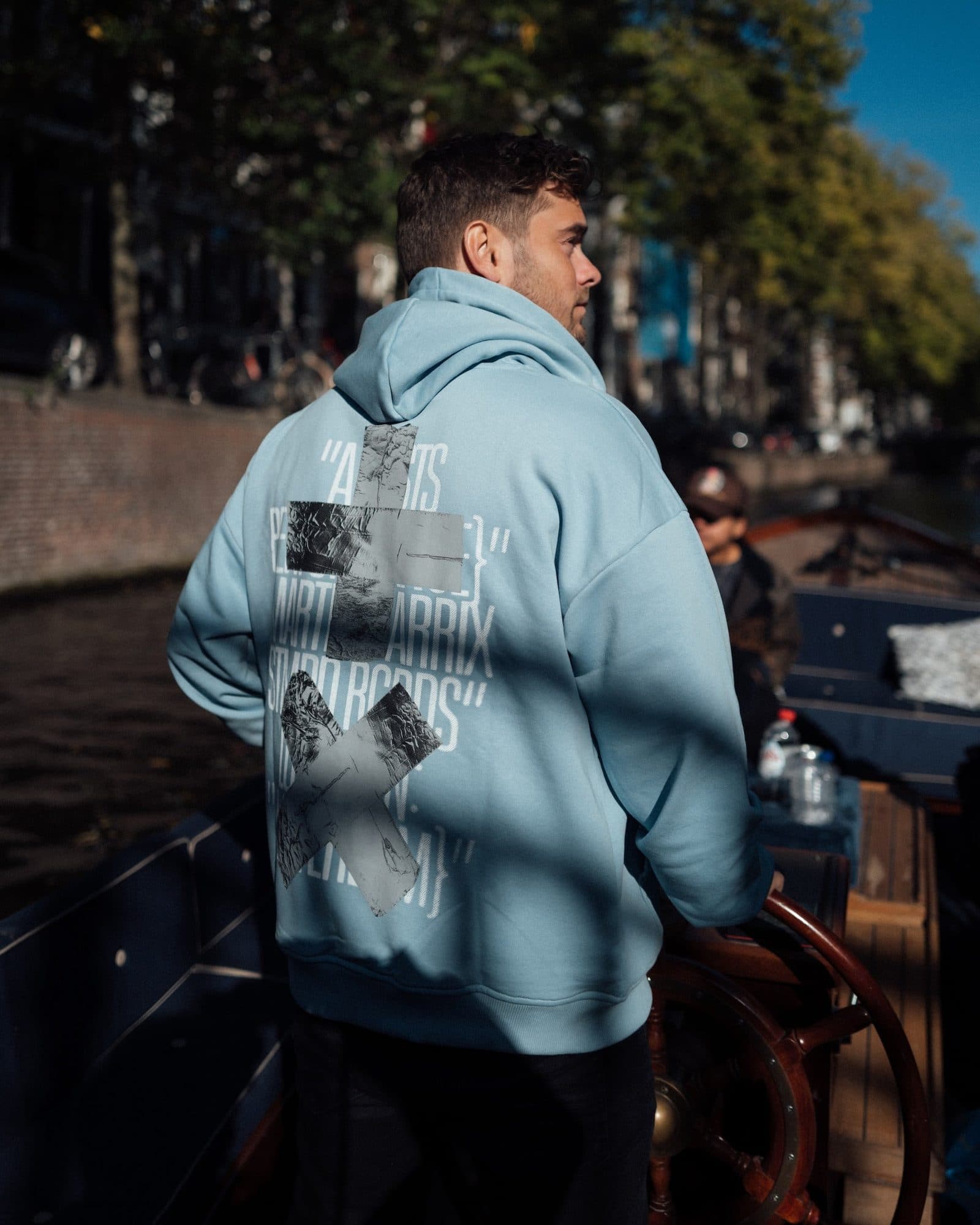 A person at the wheel of a boat on a canal, seen from behind in a light blue hoodie with a large silver cross and X over white lettering with the names Martin Garrix and STMPD RCRDS.