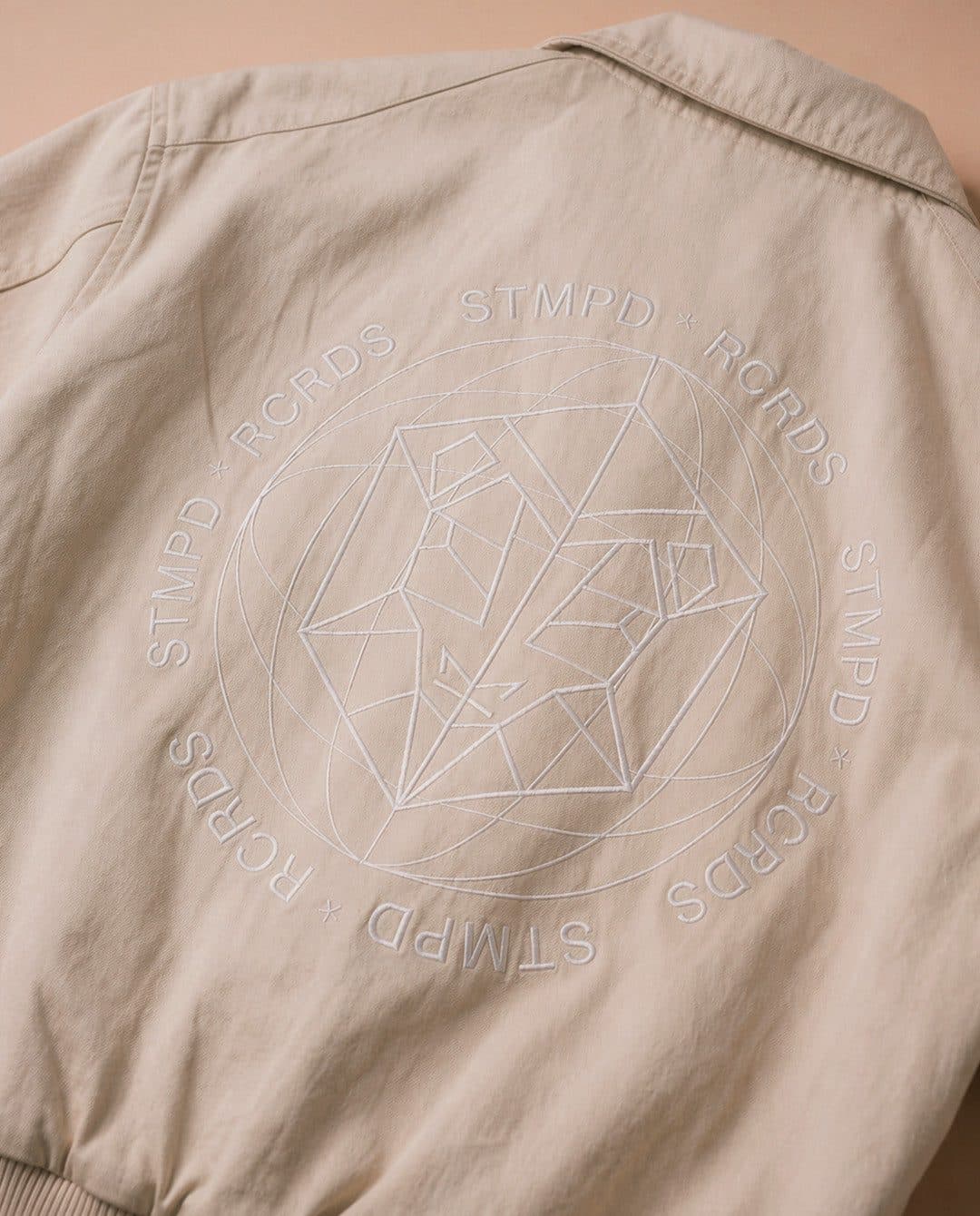 The back of a beige STMPD RCRDS summer jacket, with the label's emblem and name embroidered tone on tone.