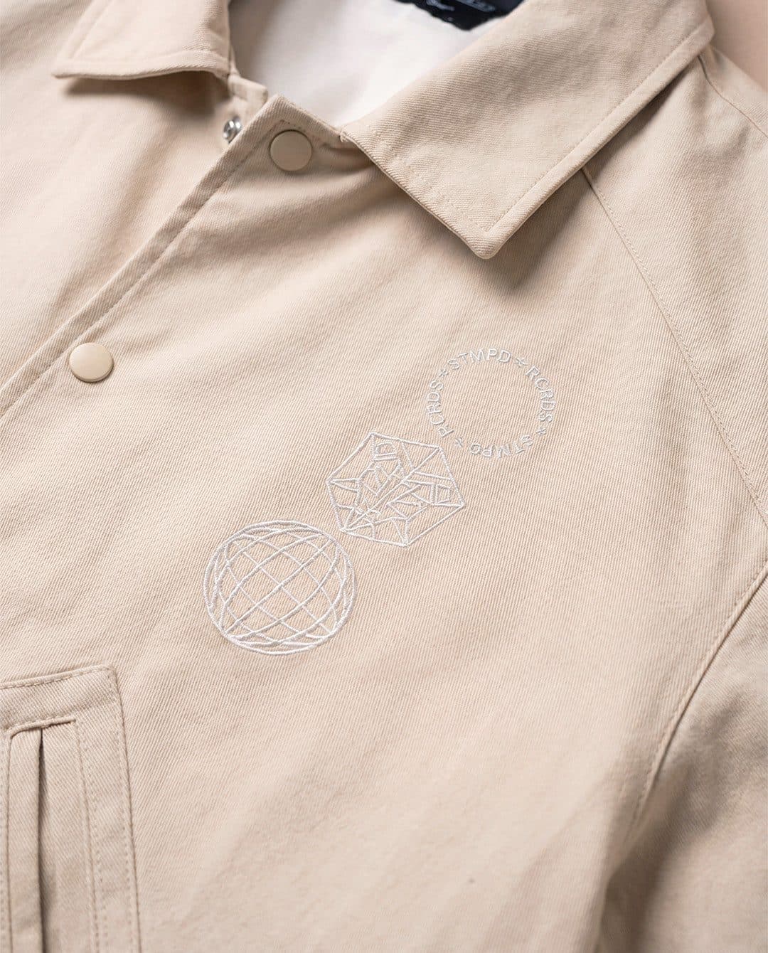 Collar and chest of a beige STMPD RCRDS summer jacket, with a globe and the label's emblem embroidered tone on tone.
