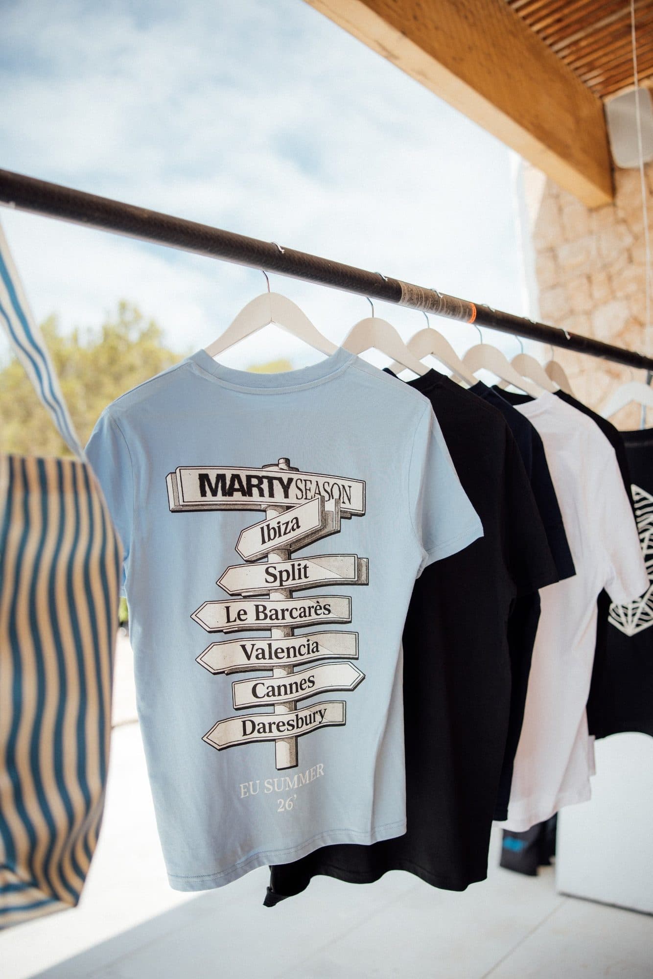 Light blue Marty Season T-shirts with a signpost print of summer destinations, on a rail outside.