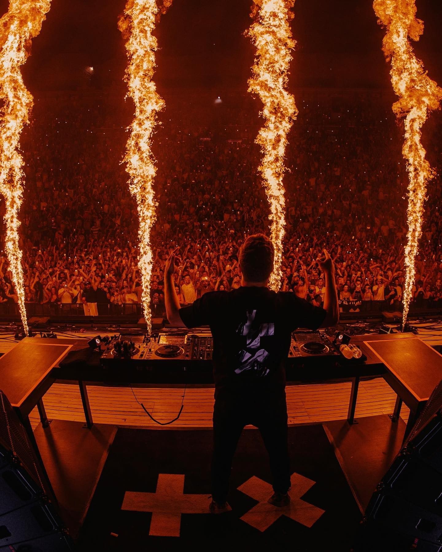 A person seen from behind at the DJ decks with both arms raised, in a black T-shirt with a cross and X back print, between four flame jets in front of a packed arena.