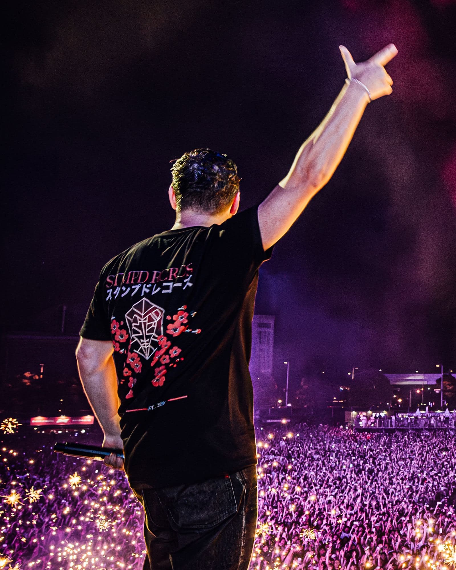 A person on a stage seen from behind, one arm raised and a microphone in hand, in a black STMPD RCRDS T-shirt with the label's name in Latin and Japanese script, its emblem and red blossoms printed on the back, above a crowd in purple light.