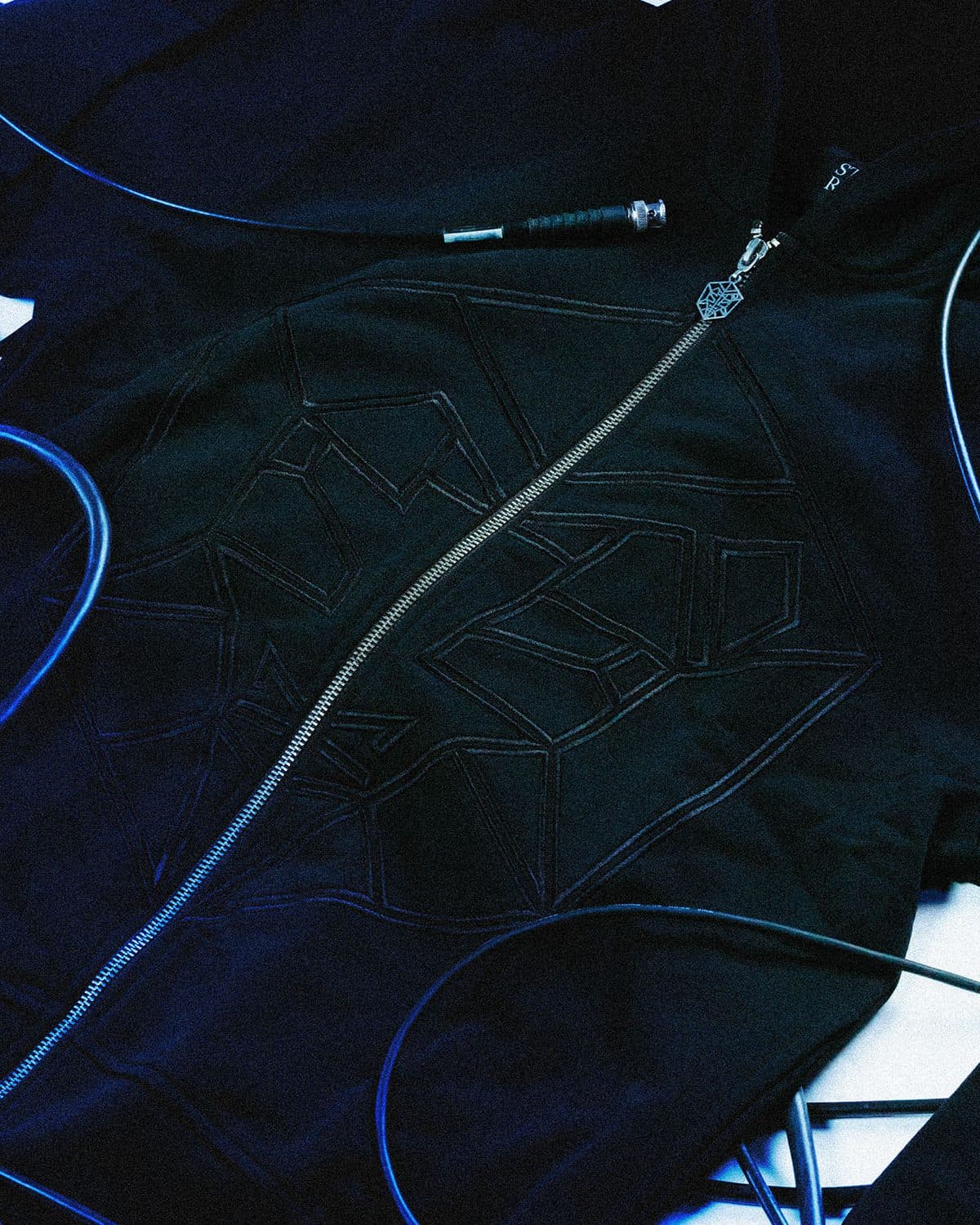 Close-up of a black STMPD RCRDS zip hoodie, with a diagonal zip, a hexagonal emblem zip pull and the label's emblem as a raised tone-on-tone outline across the chest, among dark cables.