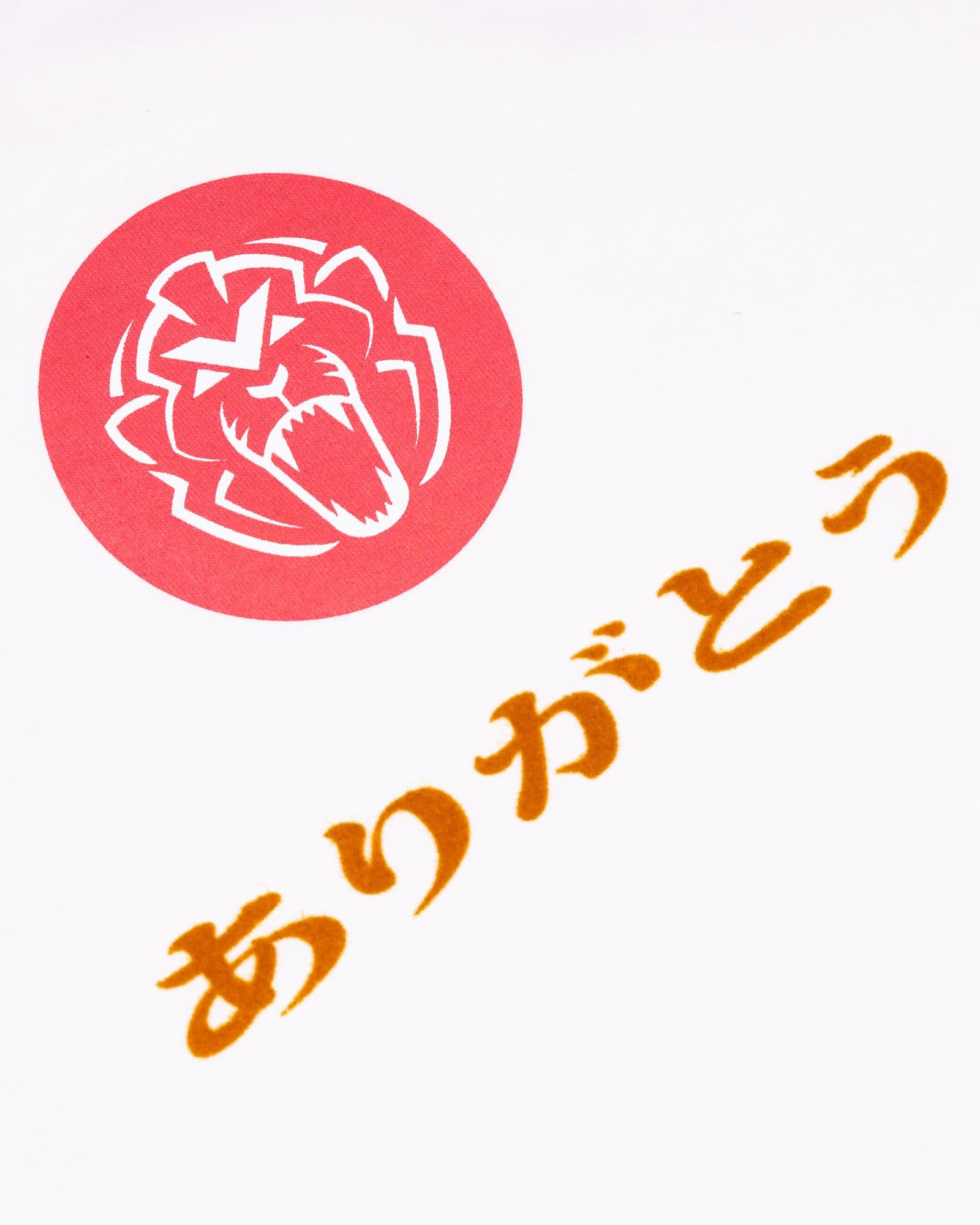 Close-up of white fabric printed with a white lion's head in a red circle, above ありがとう, Japanese for thank you, in orange.